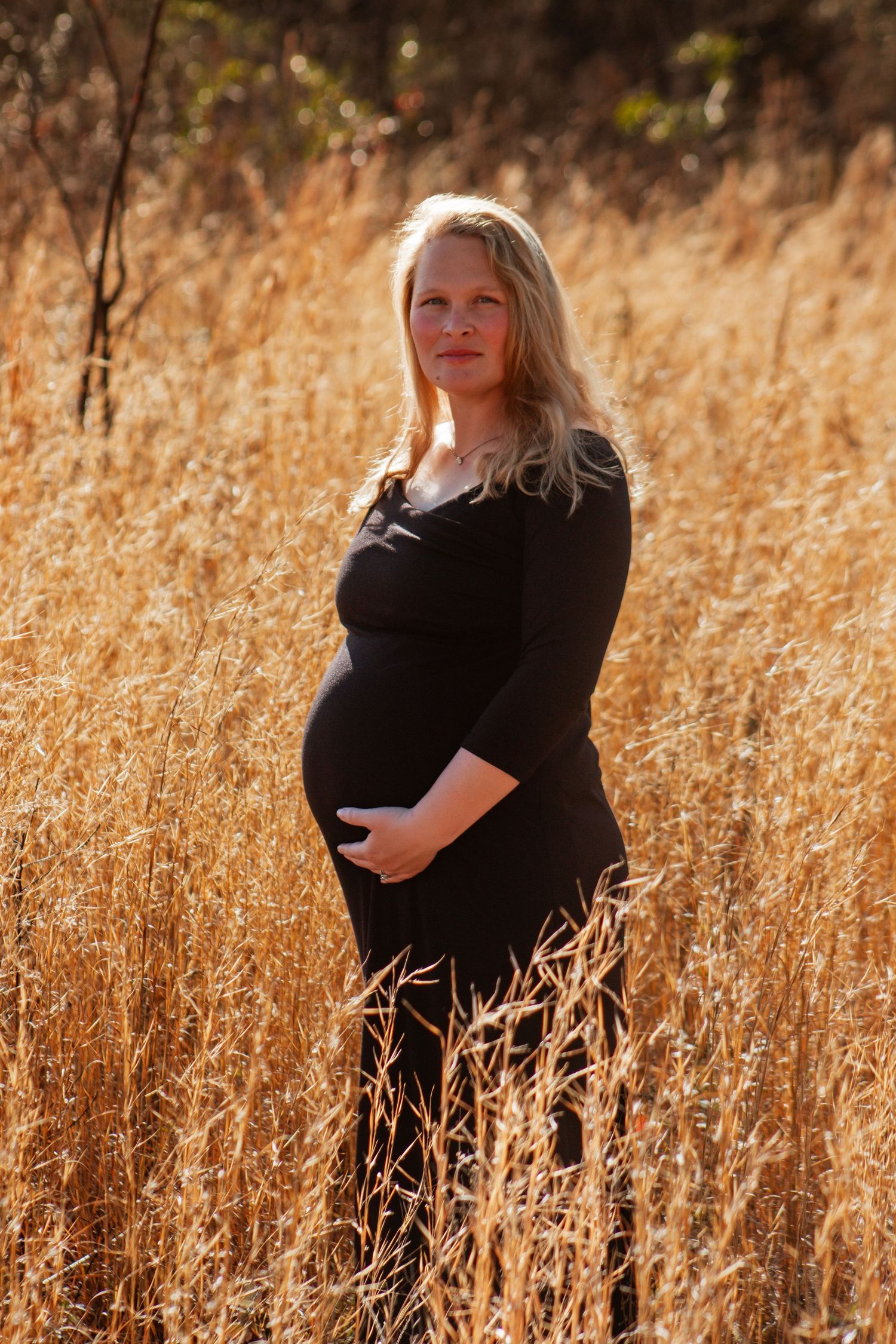 Smiling pregnant woman in black dress holding baby bump while standing in tall golden grass field outdoors.