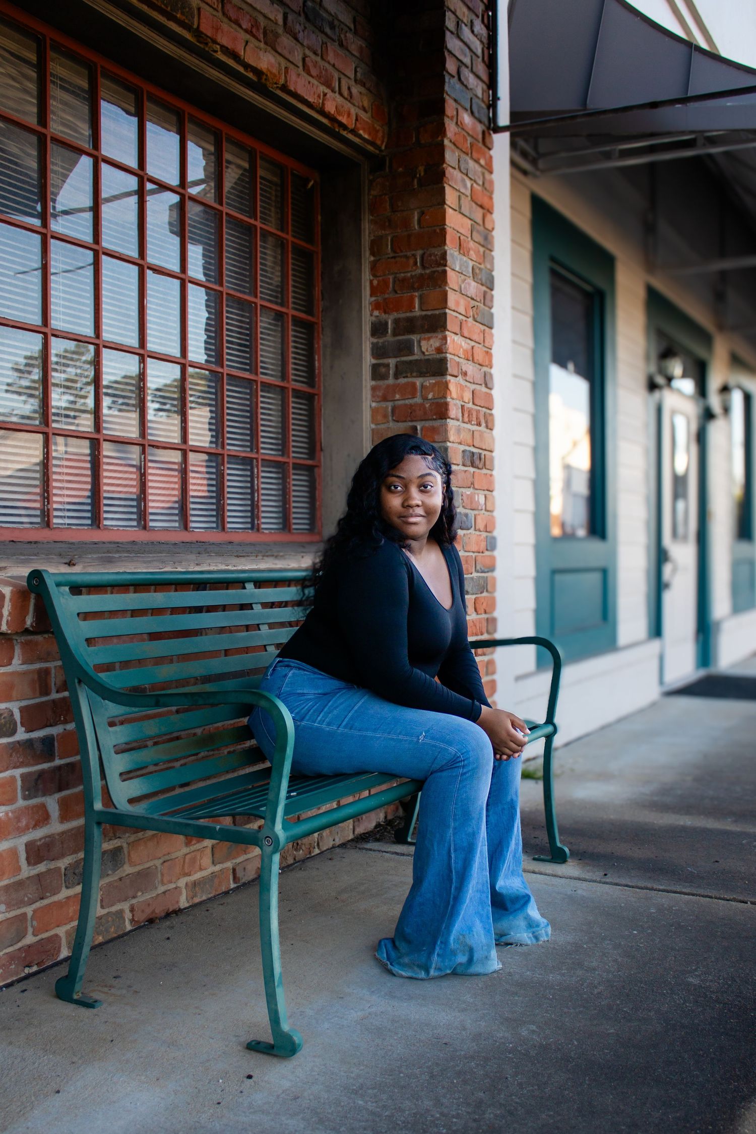 Young Black woman in black top and flare jeans sitting on teal bench outside brick building.