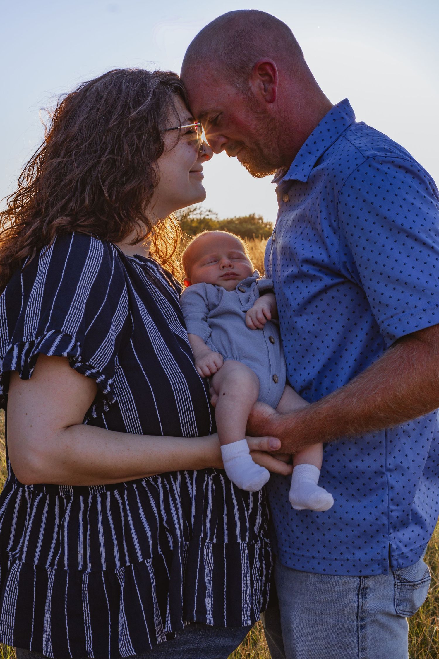 Couple touching foreheads and smiling while holding their newborn baby in a golden sunset field.