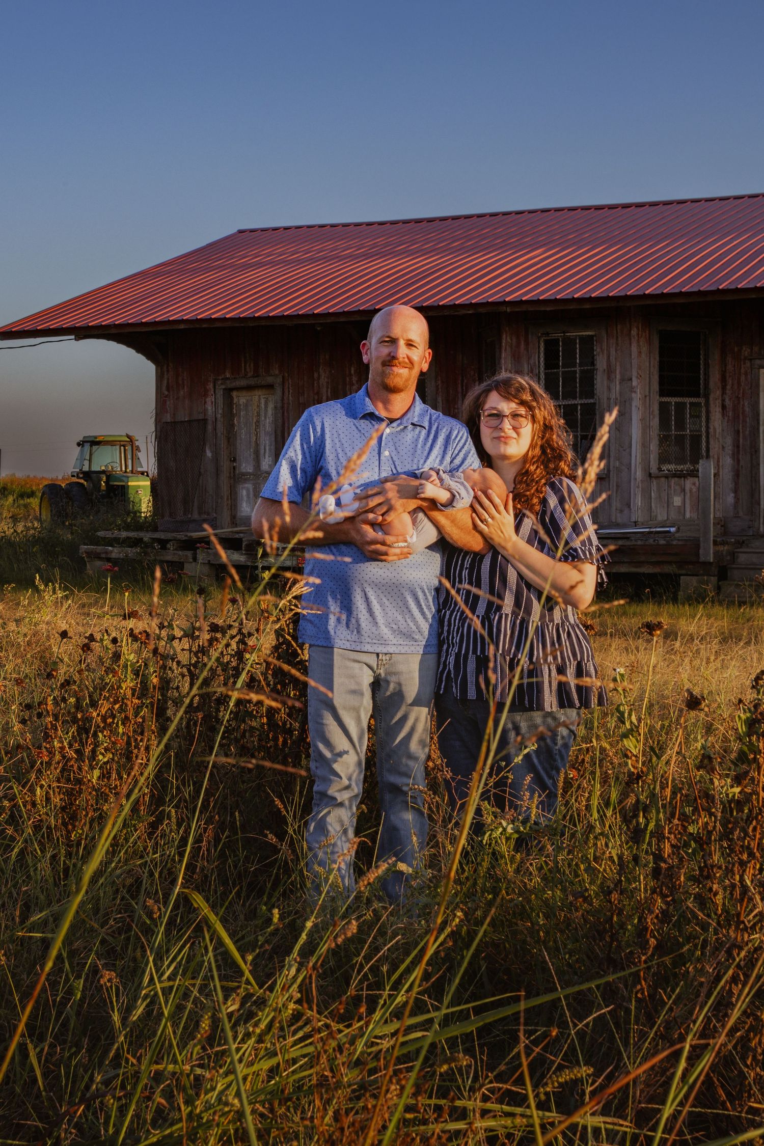 Couple holding newborn baby together standing in field near rustic red-roofed barn at golden hour.