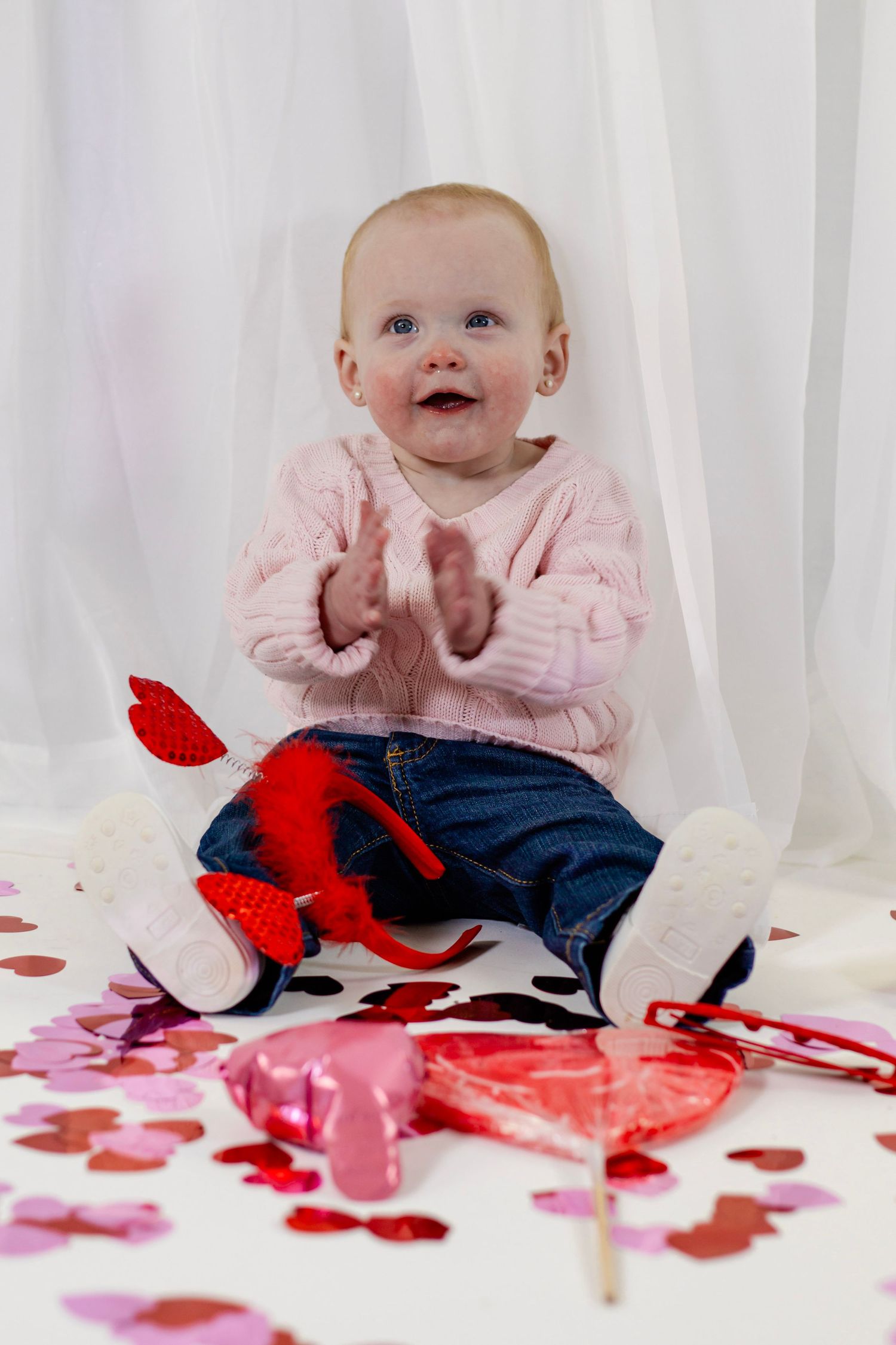 Happy baby in pink sweater clapping among Valentine's Day heart confetti, lollipops, and red stuffed animal.