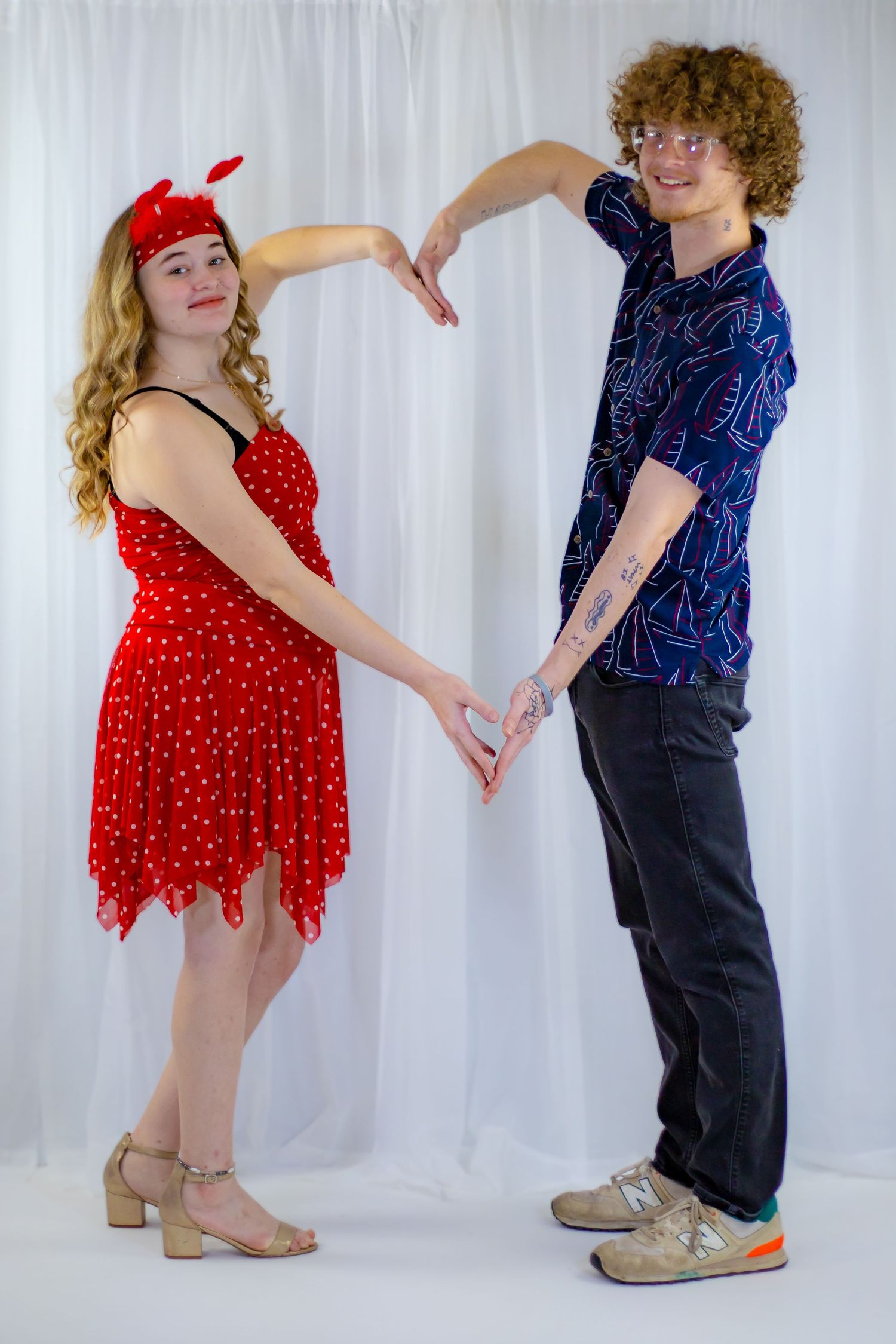 Couple forming a heart shape with their arms against a white backdrop in Valentine's Day outfits.