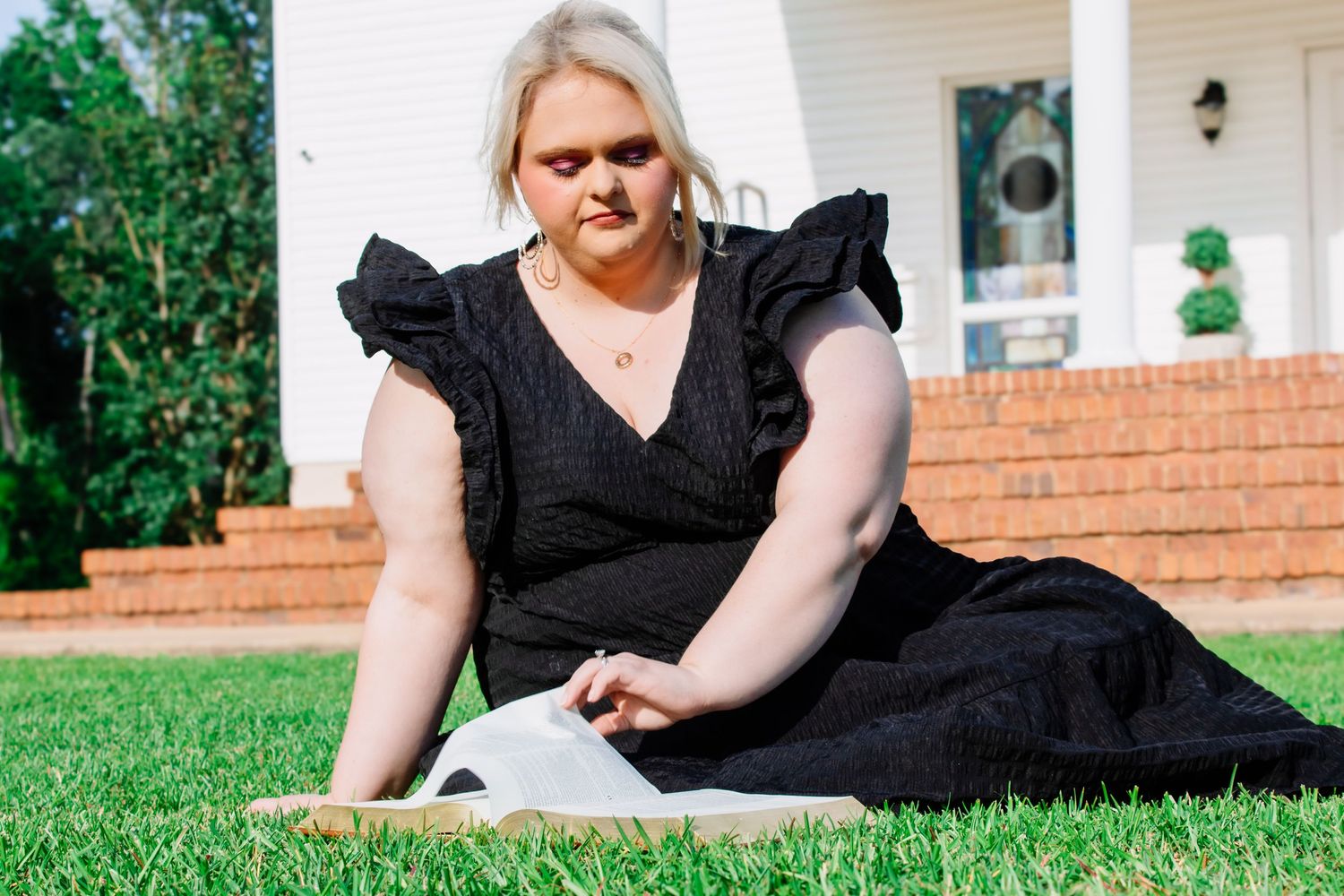 Blonde woman in black ruffled dress sitting on green grass outdoors holding an open book.