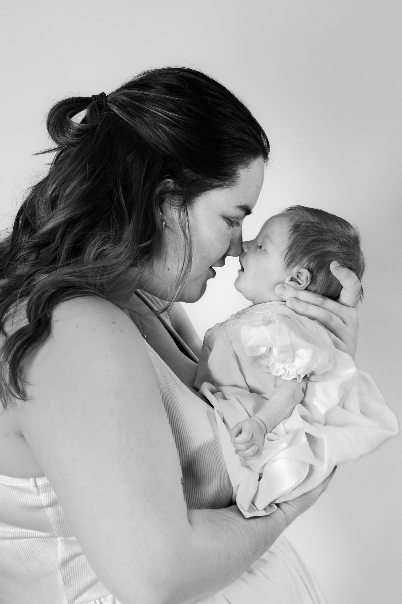 Black and white photo of a mother tenderly touching noses with her newborn baby wrapped in a soft blanket.