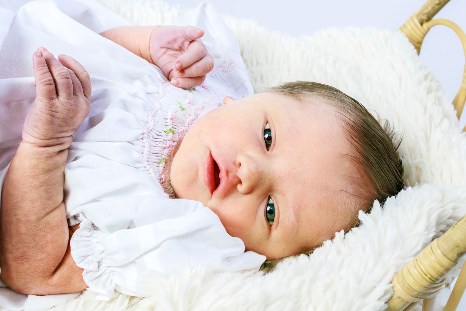 Adorable newborn baby girl with blue eyes lying in a fluffy white wicker basket wearing a white outfit.