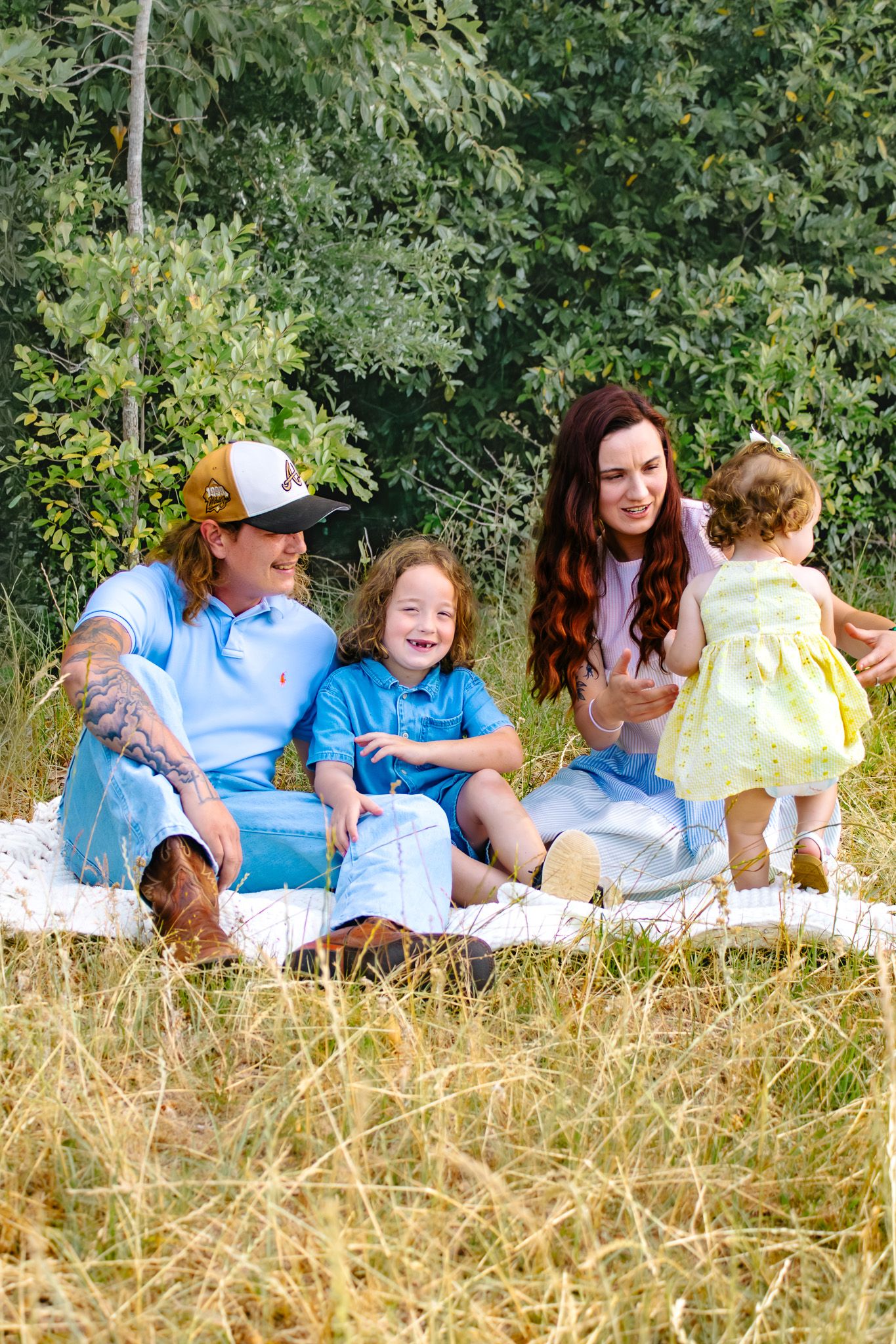 Family of four enjoying a outdoor picnic in a grassy field surrounded by lush green trees.