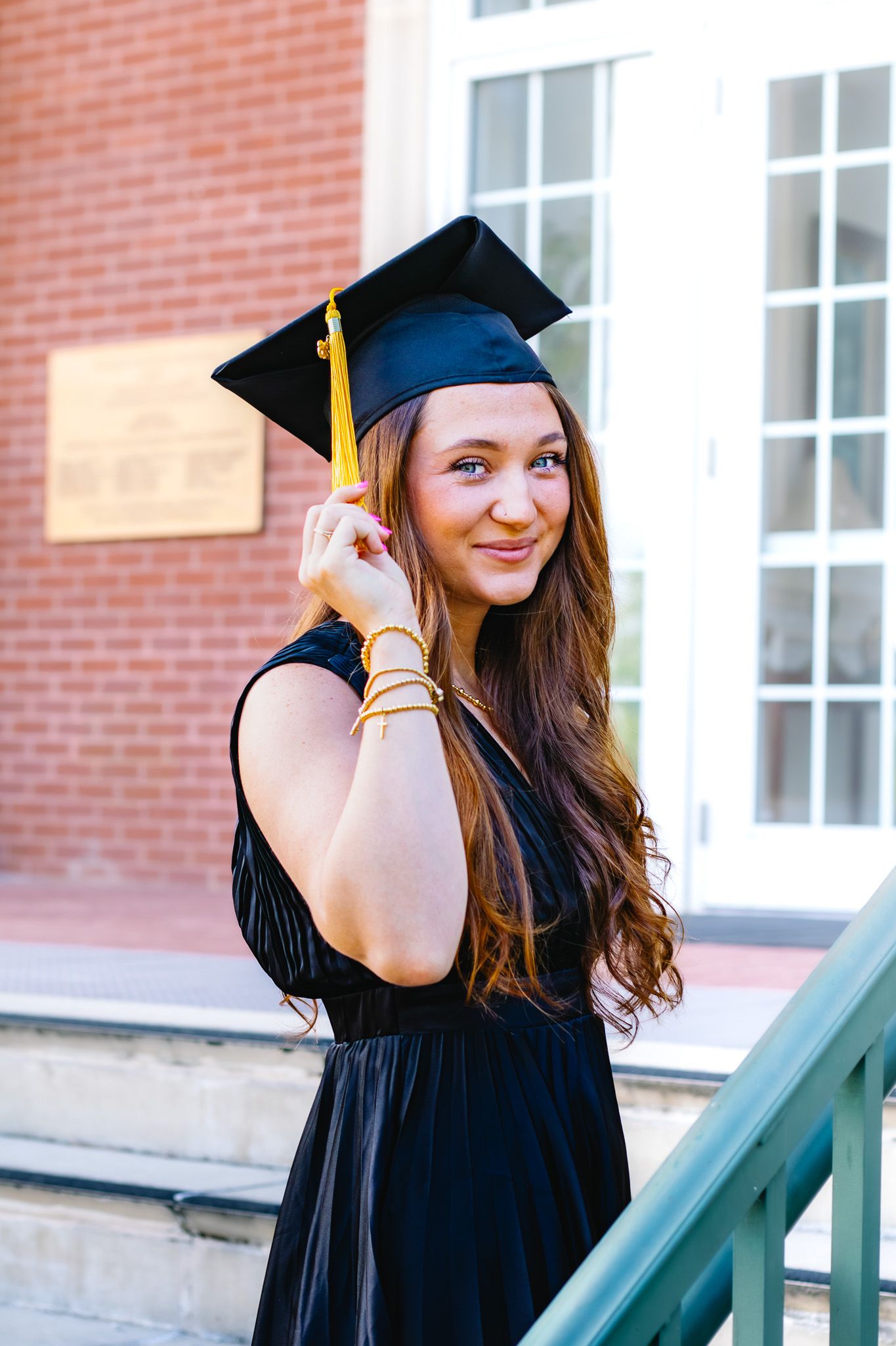 Smiling graduate in black cap and gown holds tassel beside brick building at campus.