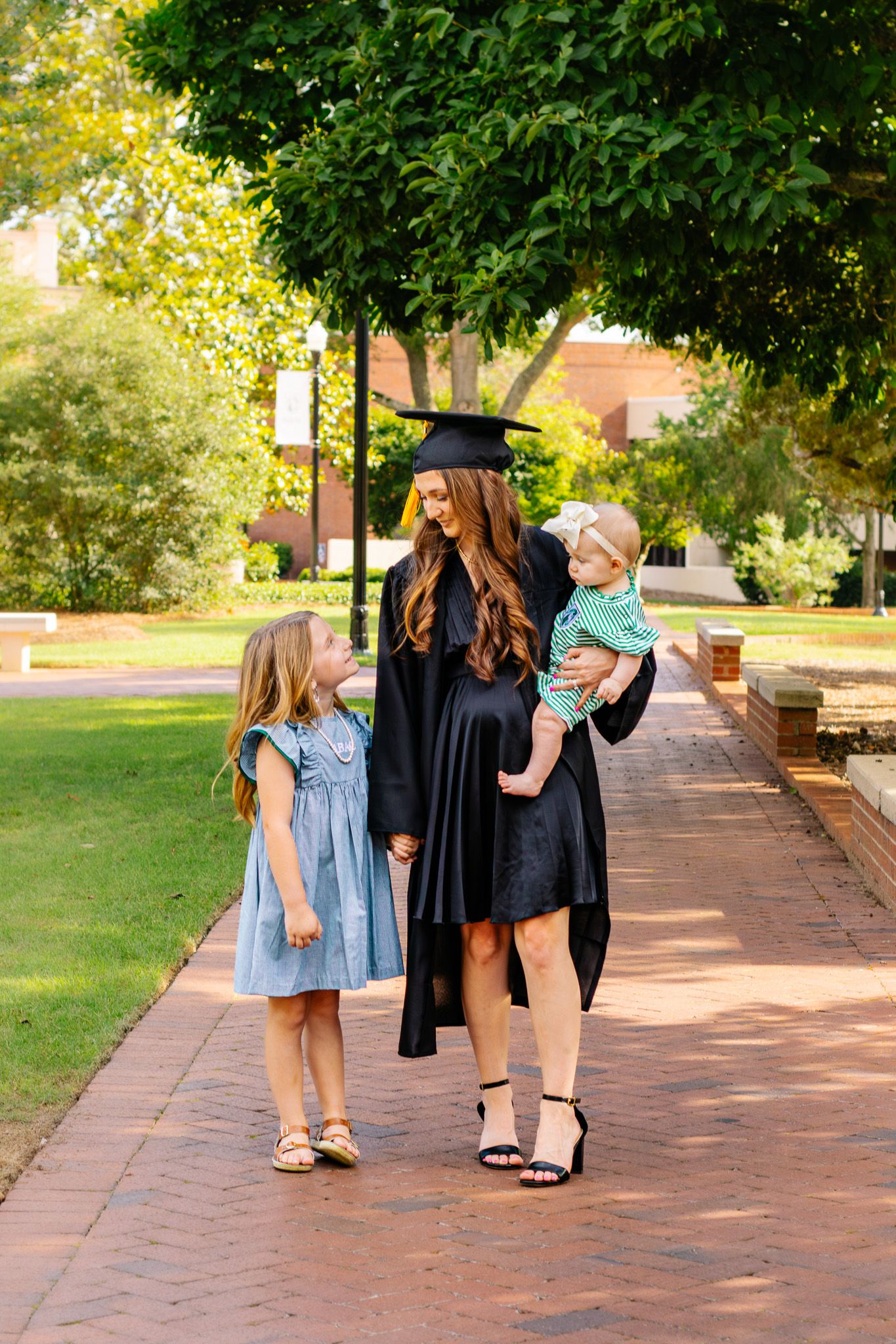A woman in graduation cap and gown holds a baby while talking with a young girl on a brick campus walkway.