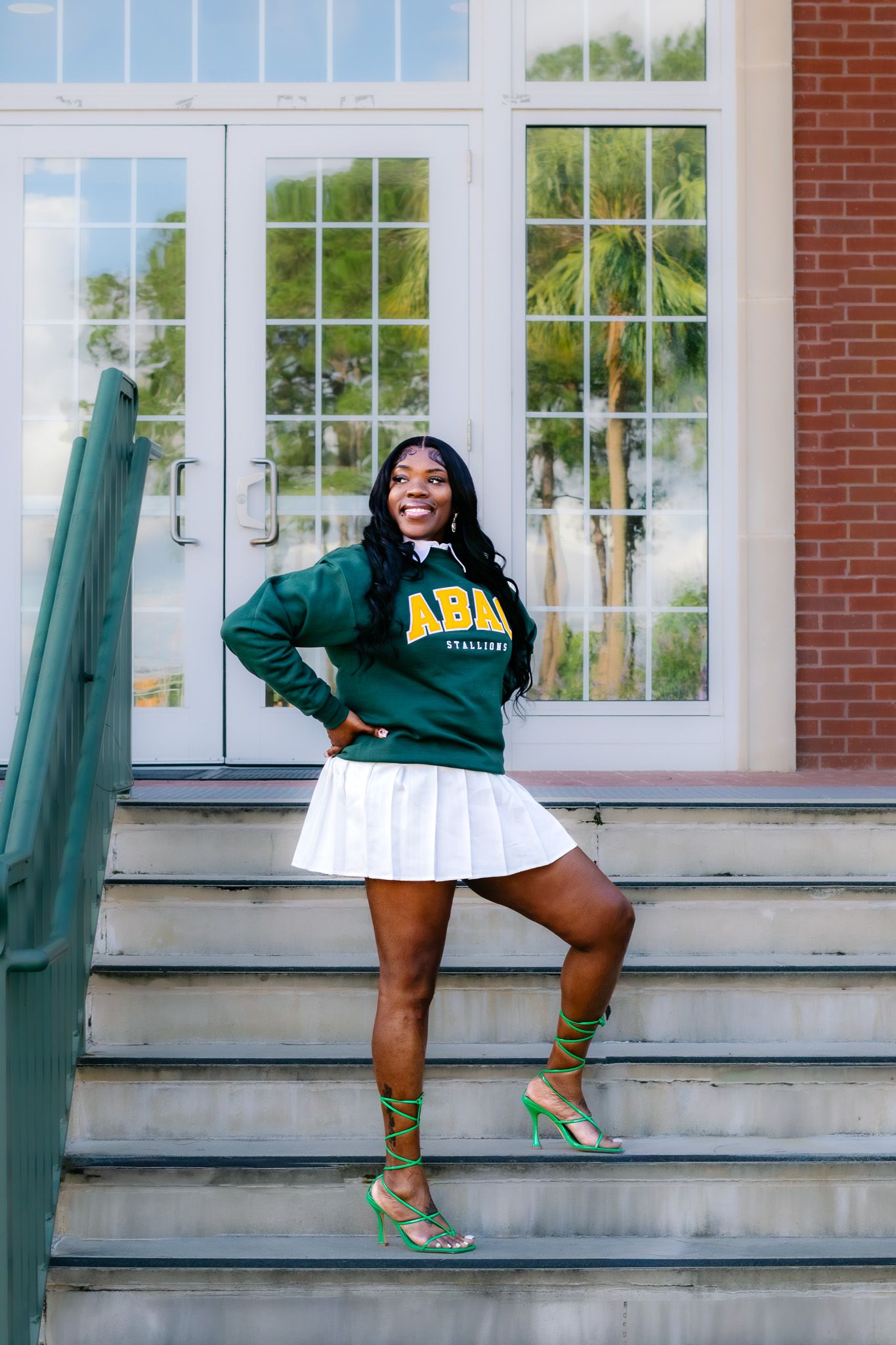 Smiling woman in green ABAU sweatshirt, white pleated skirt, and green heels posing on steps outside a brick building.