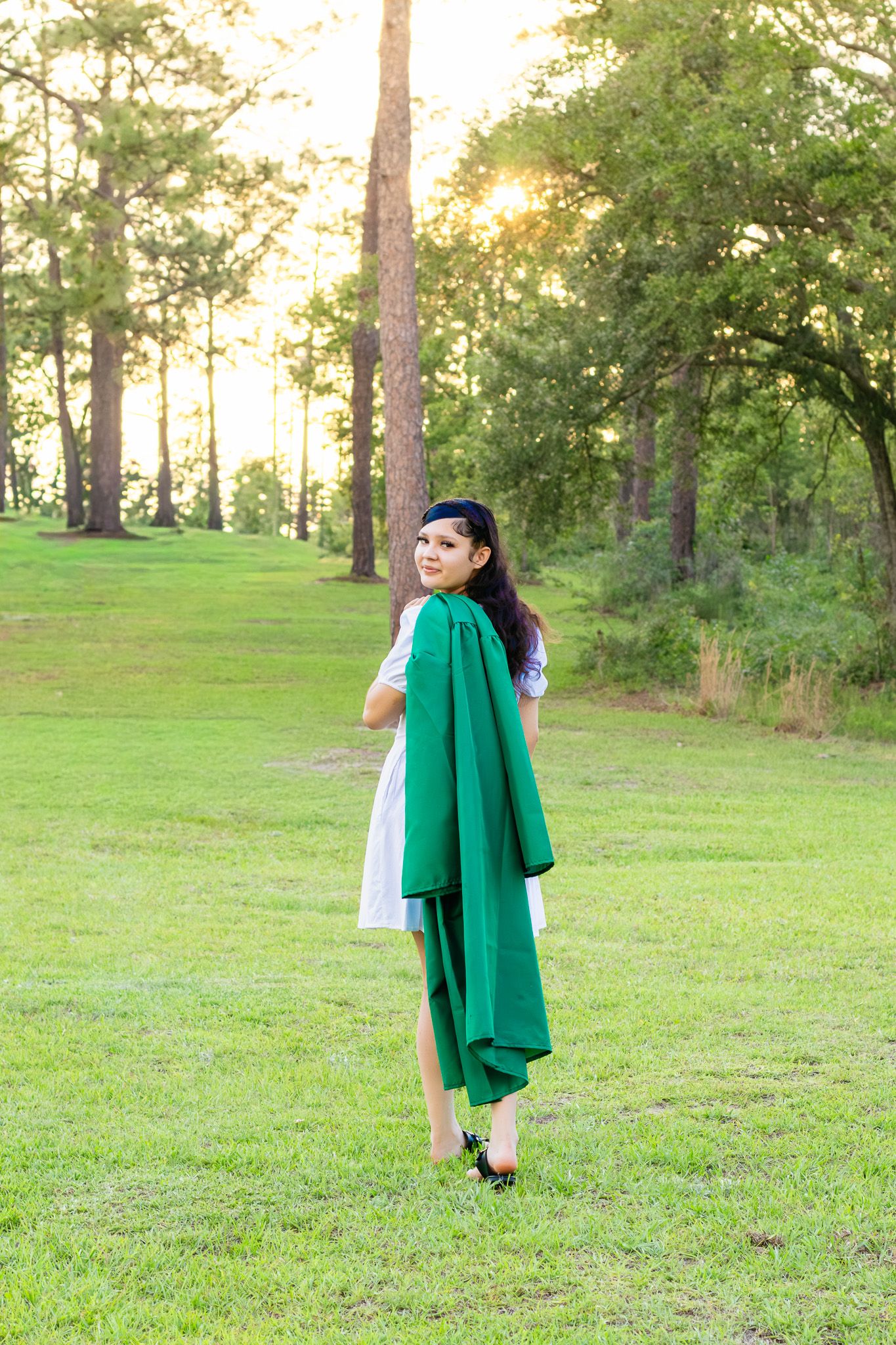 Young woman in white dress holding green graduation gown stands on lush grass at sunset among tall pine trees.
