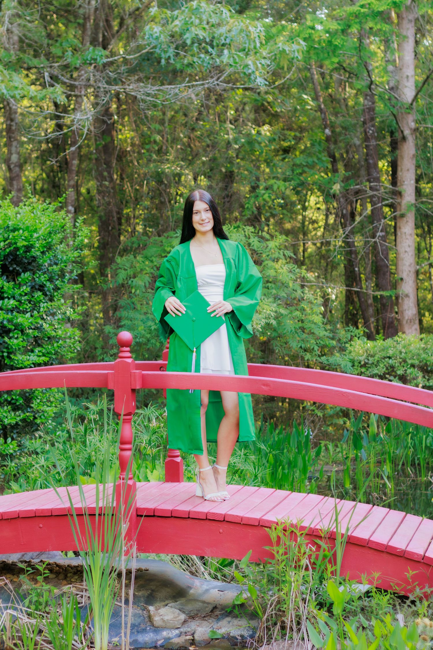Graduate in green gown holding cap while standing on red arched bridge in garden.