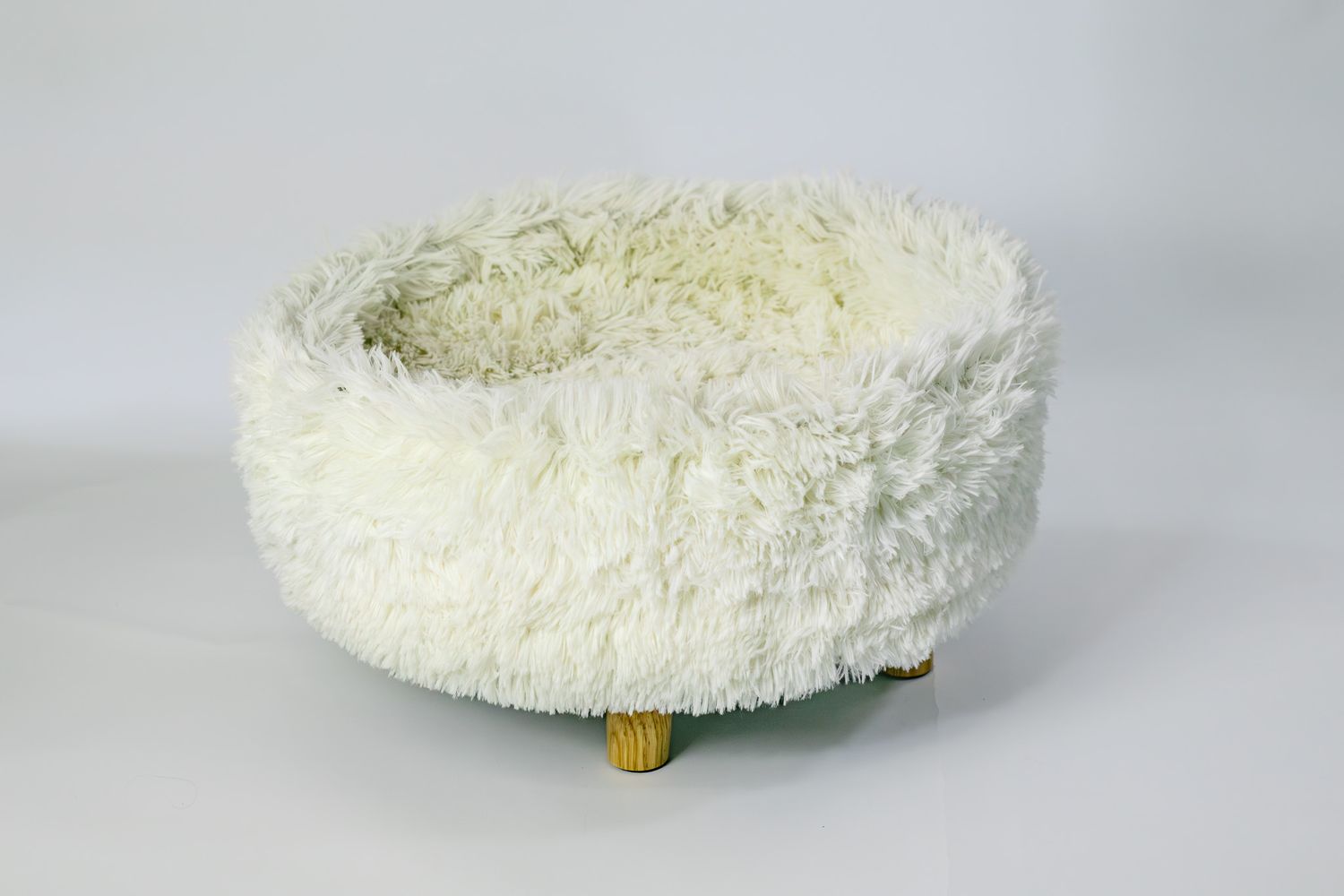 White fluffy round pet bed with soft faux fur exterior and small wooden legs, ideal for cats or small dogs.