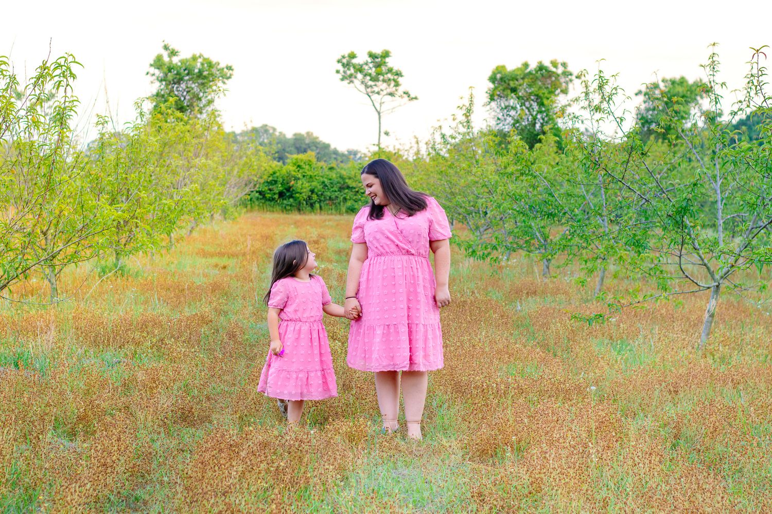 A mother and daughter in matching pink dresses hold hands while walking through a meadow.