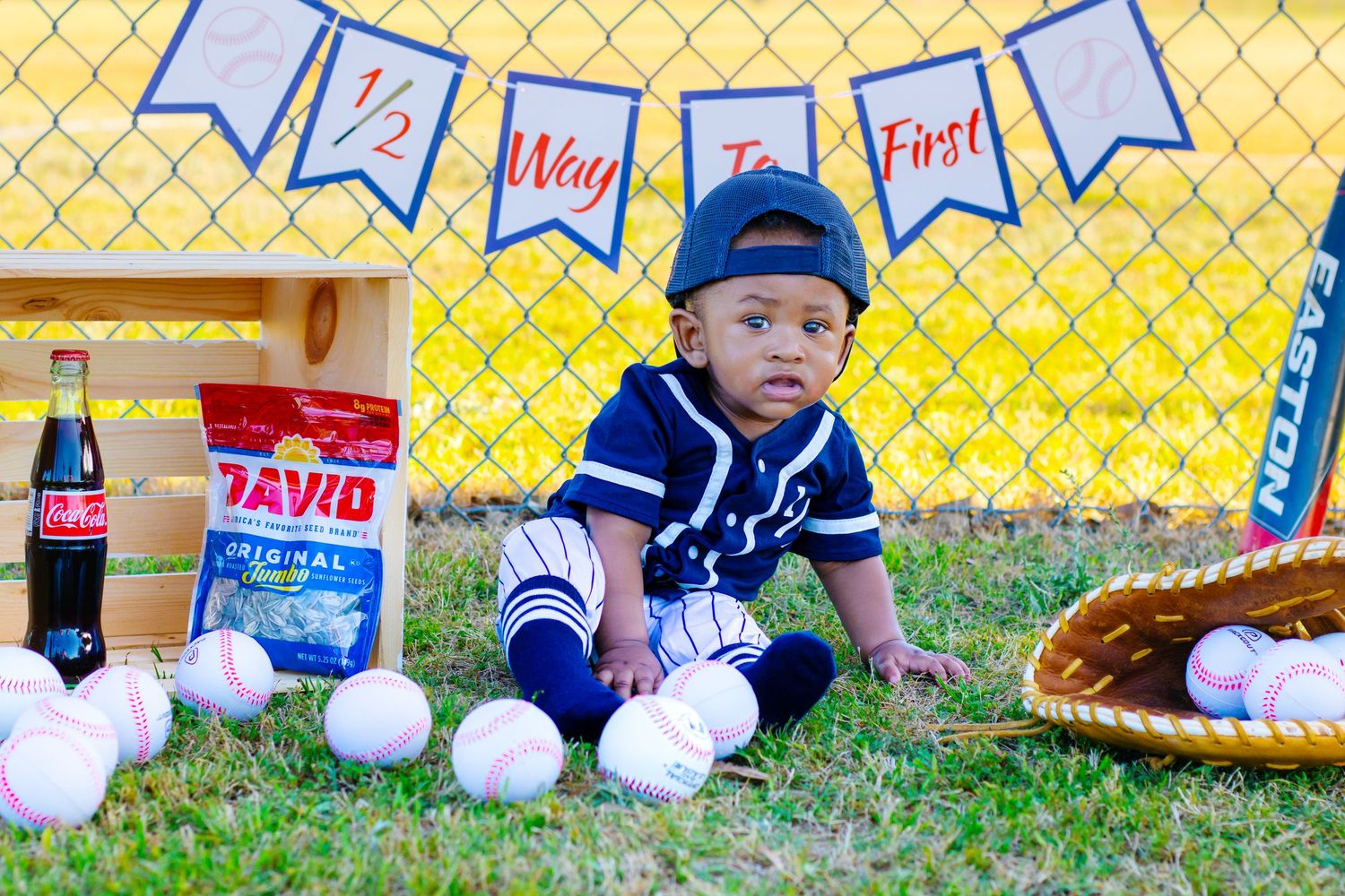 Baby in baseball uniform sits on grass with baseballs, glove, and snacks, celebrating half birthday milestone.