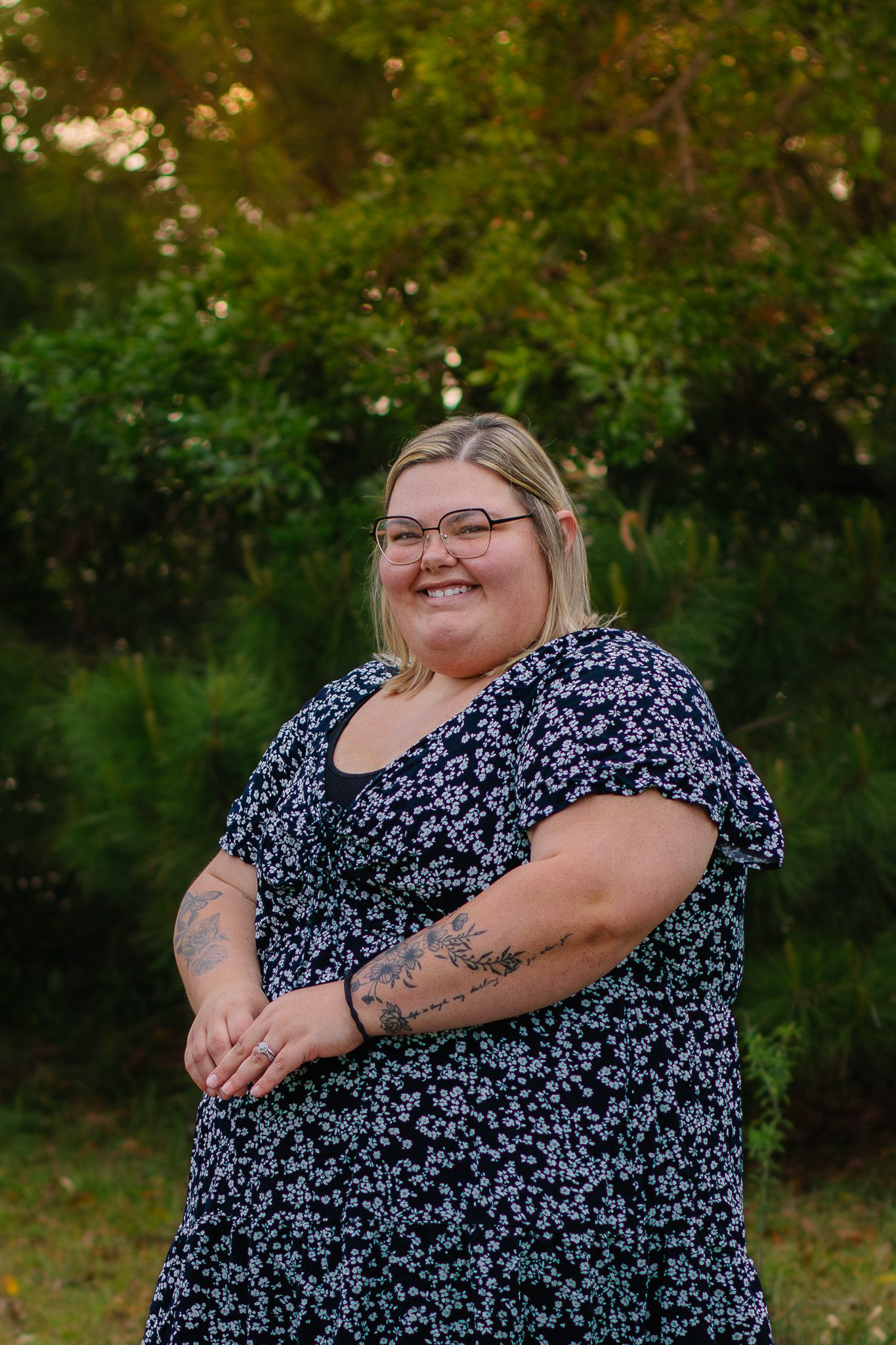 Smiling blonde woman in floral dress with glasses and tattoo posing outdoors.