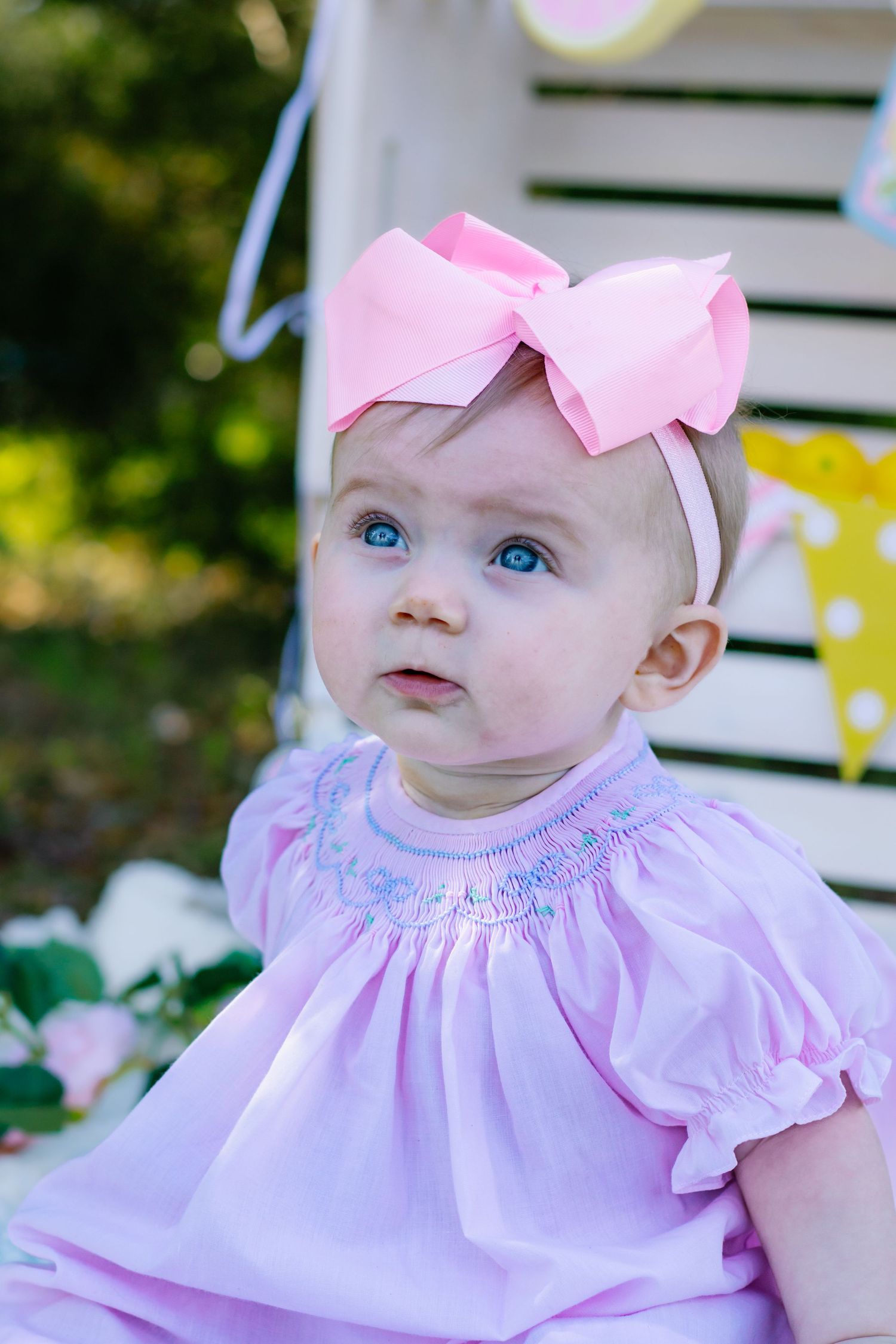 Blue-eyed baby girl in pink smocked dress with large pink bow headband looking up curiously.