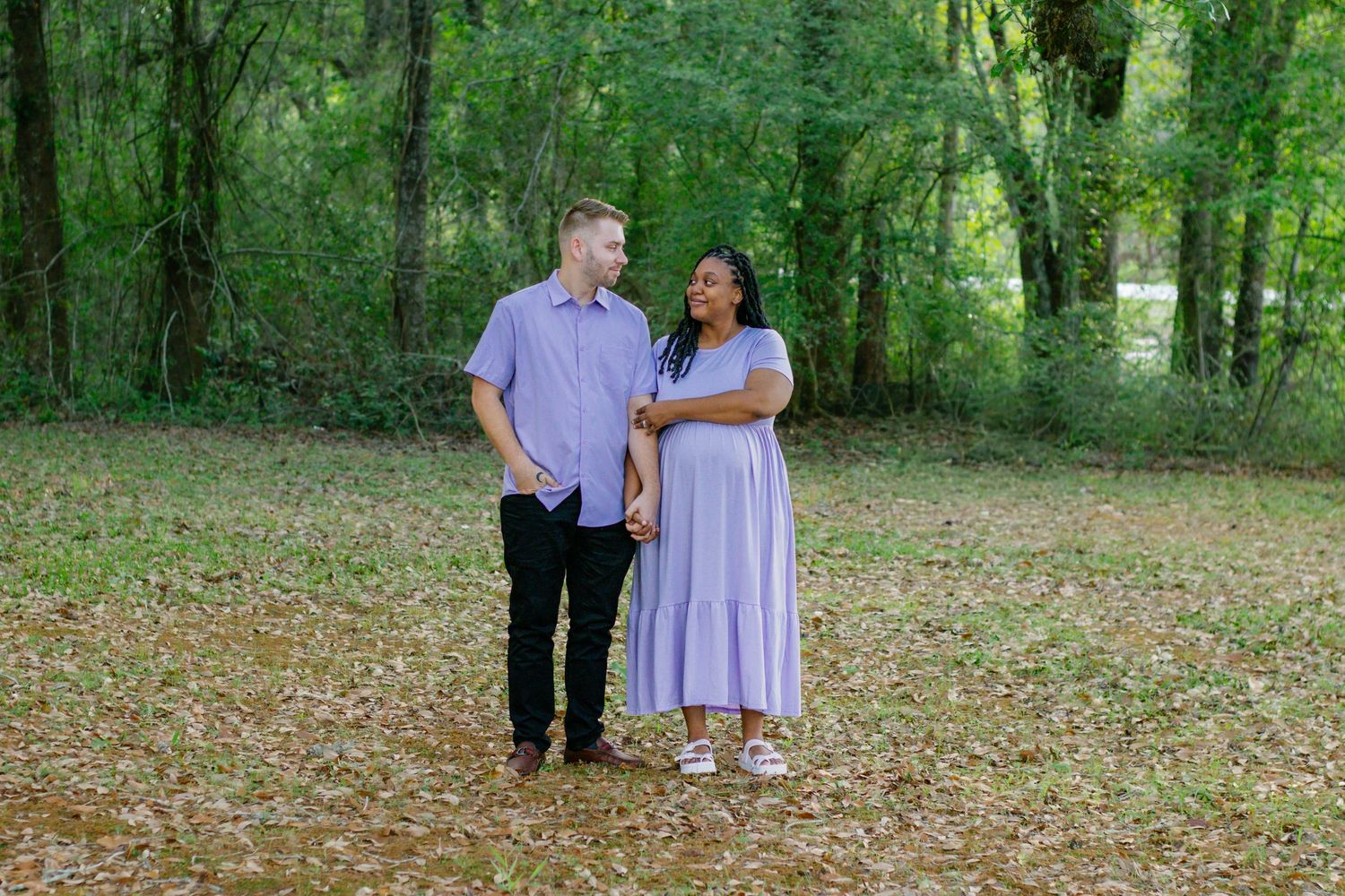 A couple in matching lavender outfits holds hands while smiling at each other in a lush green forest clearing.