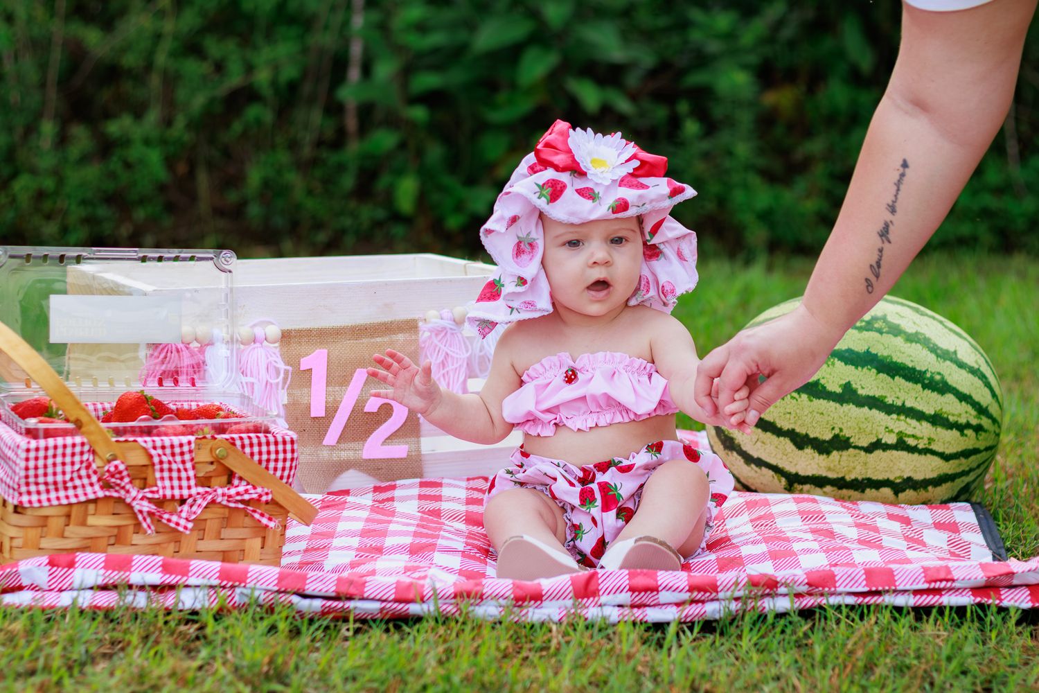 Sweet baby in strawberry romper and bow hat poses with watermelon on red gingham blanket for 1/2 birthday.