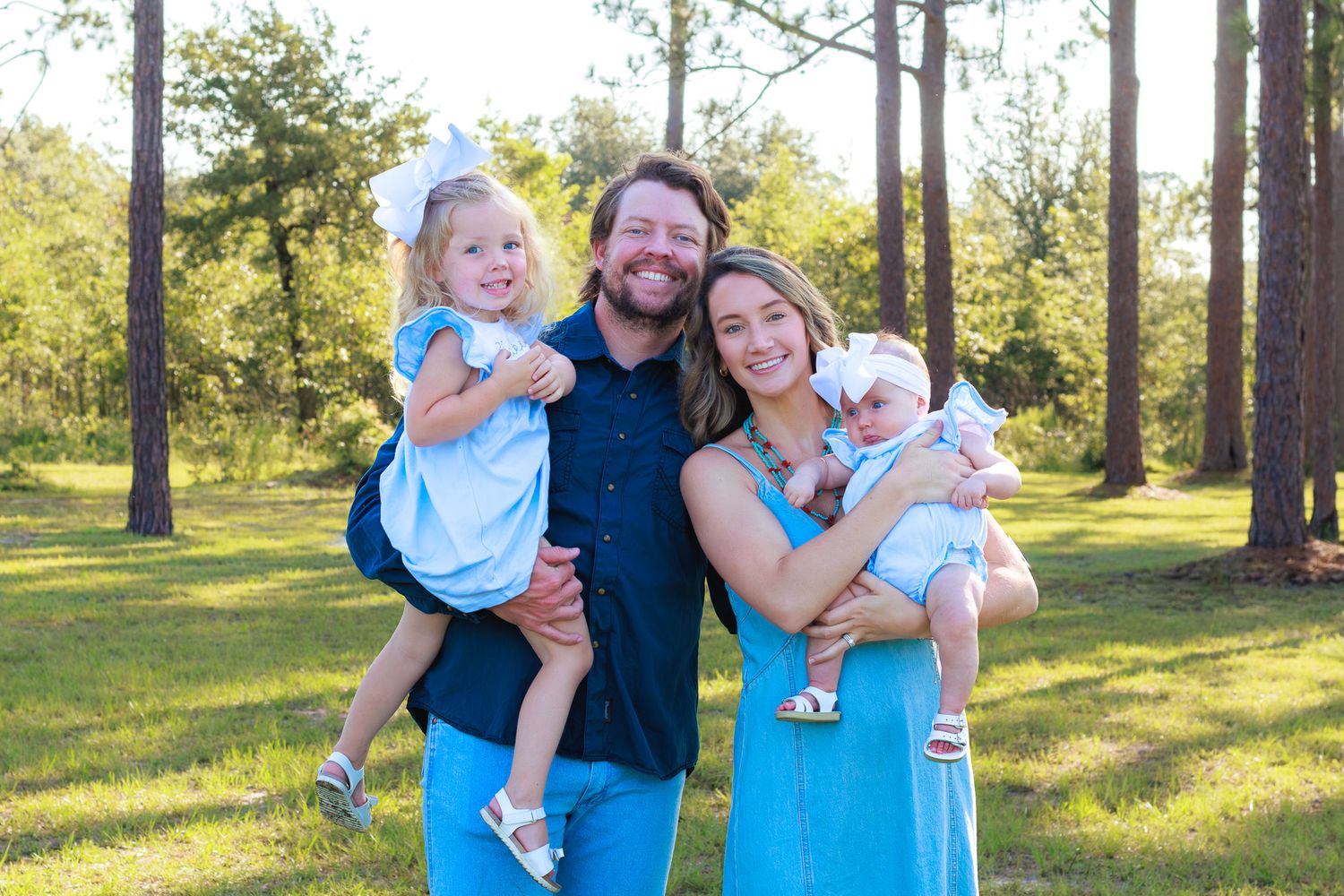 Happy family of four posing outdoors in matching blue outfits, parents holding two young daughters among tall pine trees.