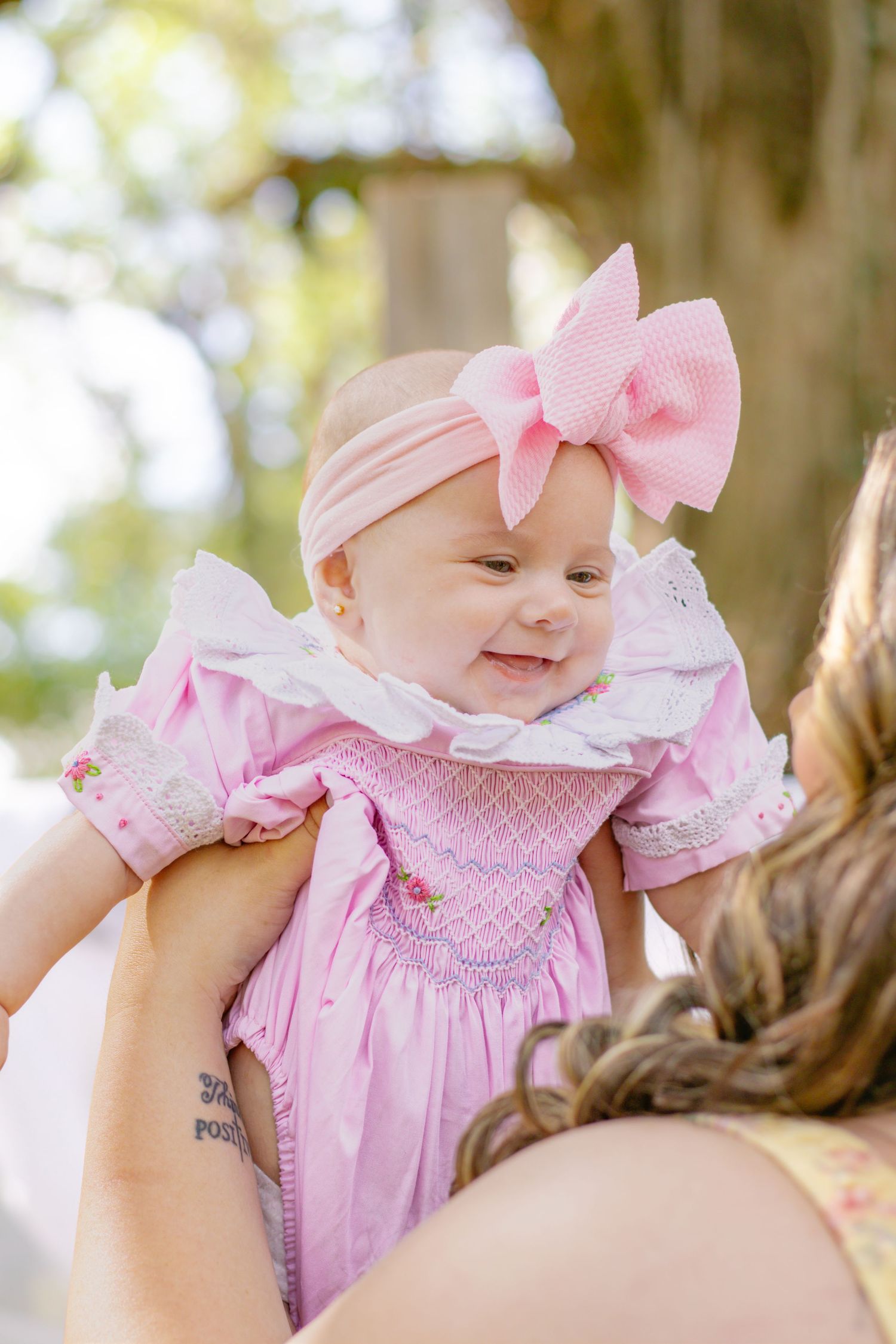 Joyful baby girl in pink smocked dress with bow headband being lifted by mother outdoors.