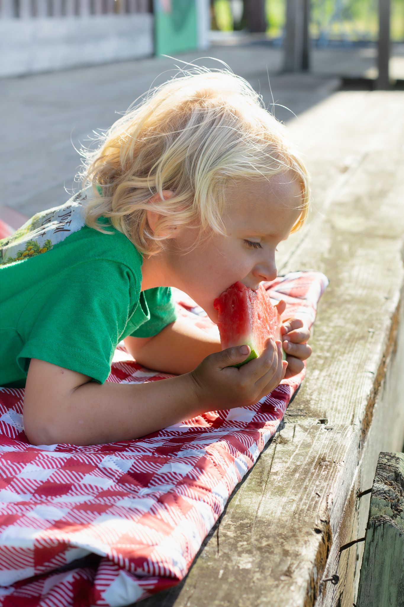 Blond child lying on red checkered cloth outdoors eating a fresh slice of watermelon on a sunny day.