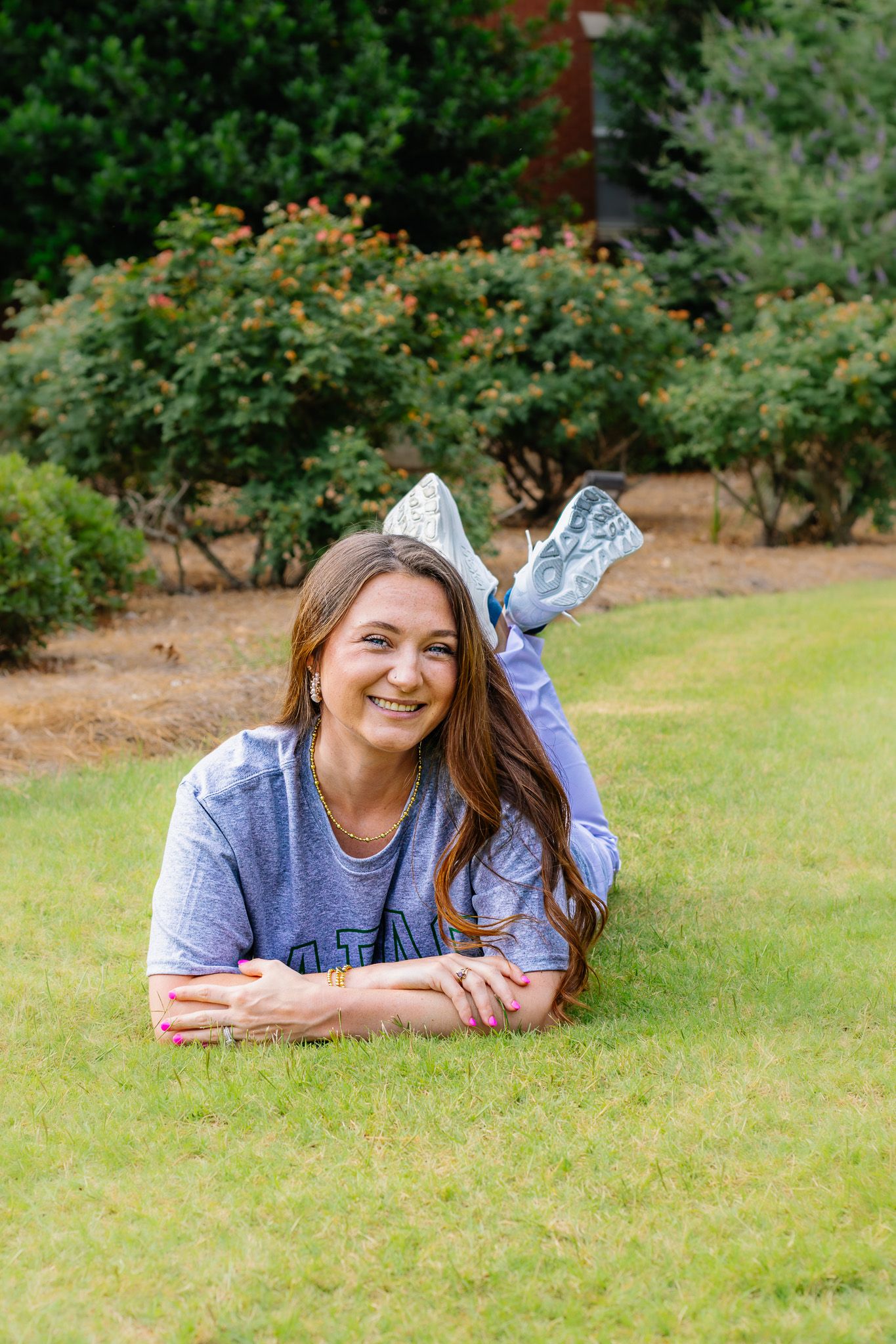 Happy young woman in college t-shirt lies on green grass smiling with feet kicked up behind her.