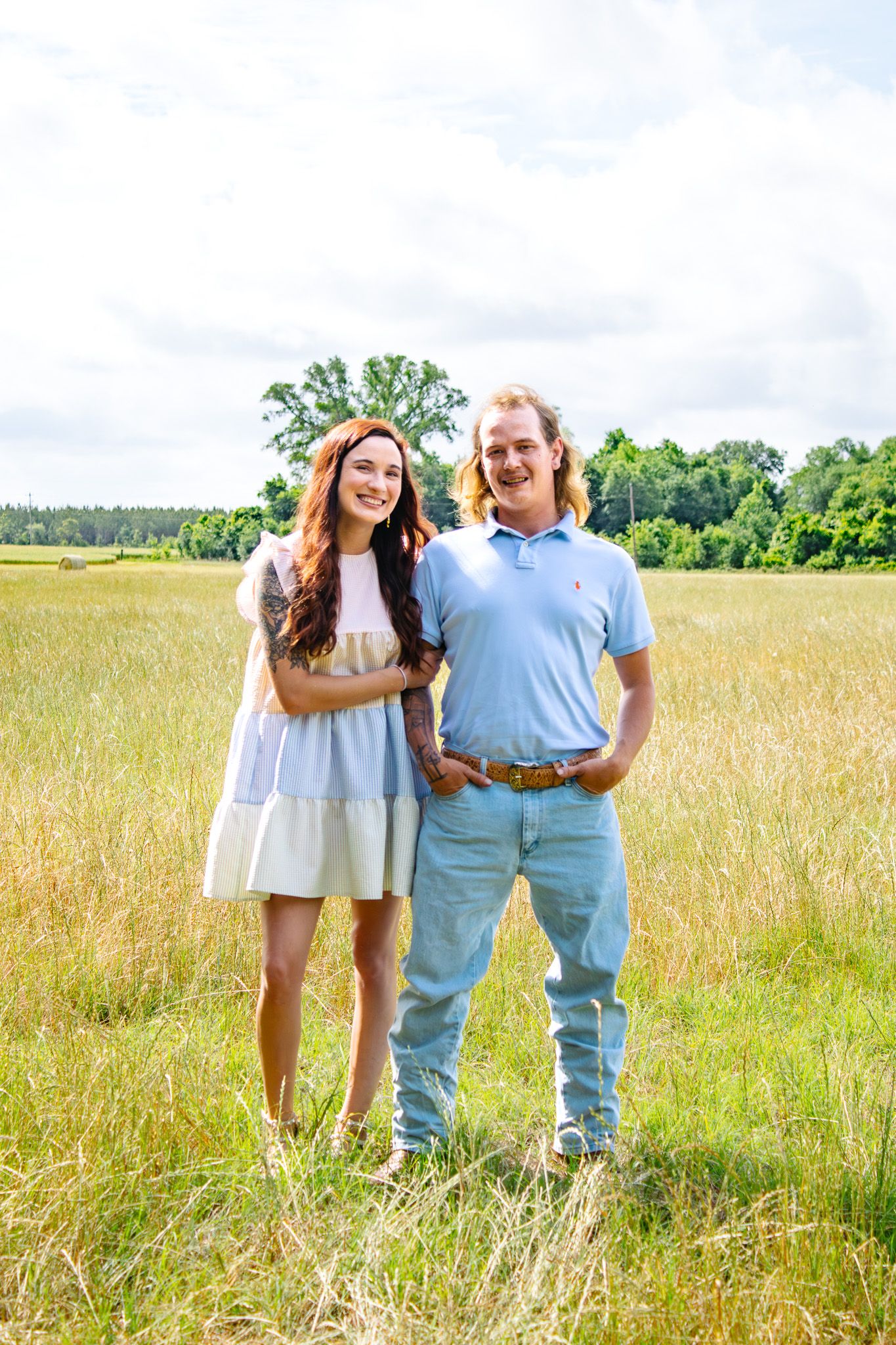 Smiling couple standing together in a sunny golden field with green trees and clouds in the background.