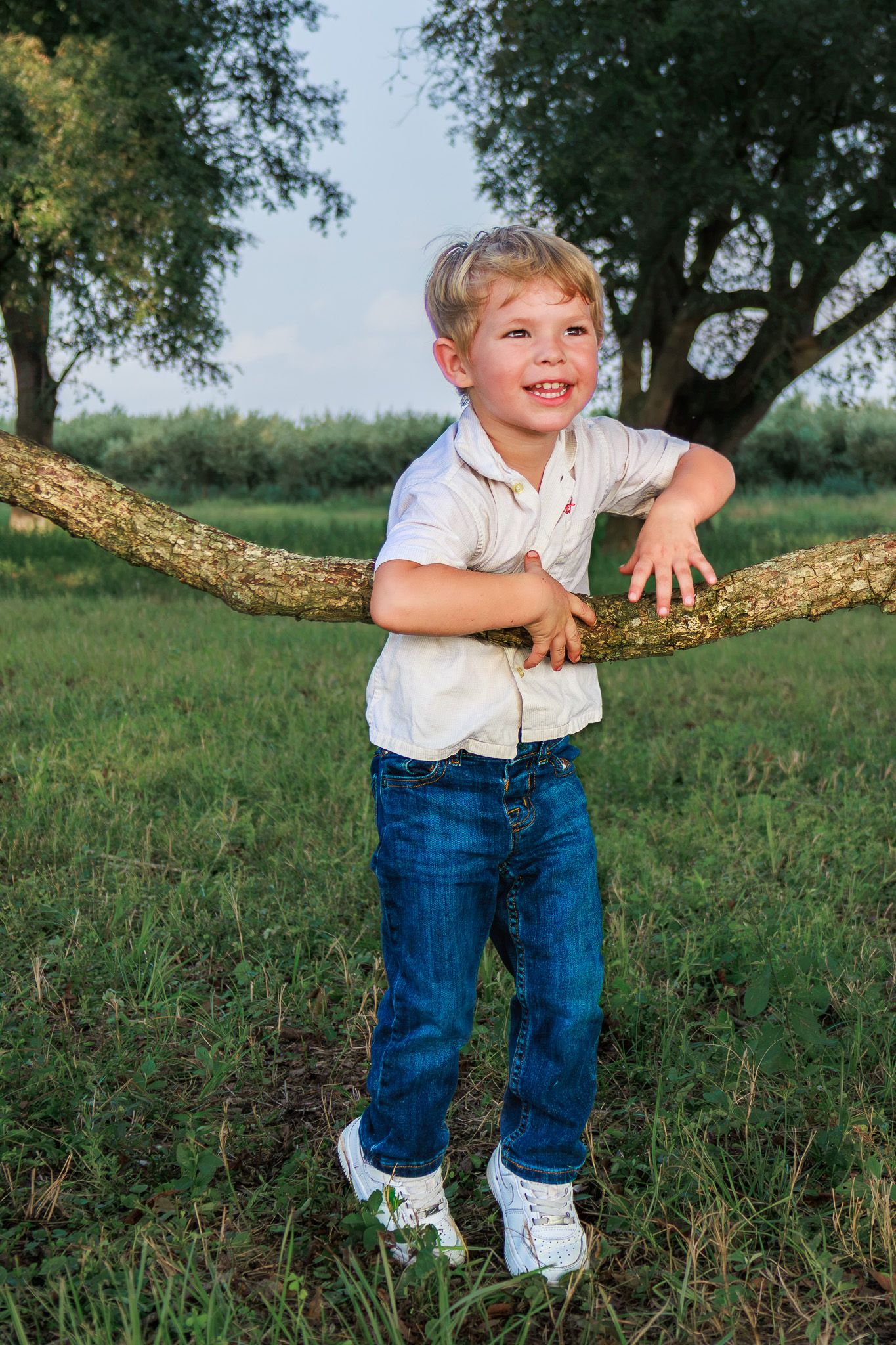 A cheerful young boy in a white shirt and jeans leans on a low tree branch in a lush green outdoor setting.