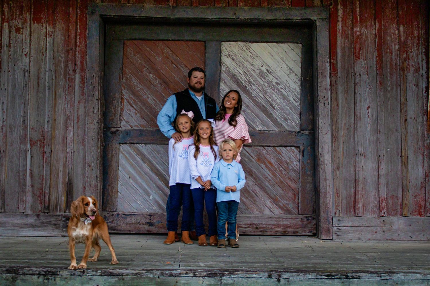 Family of five with a Spaniel dog posing before a rustic red barn door.
