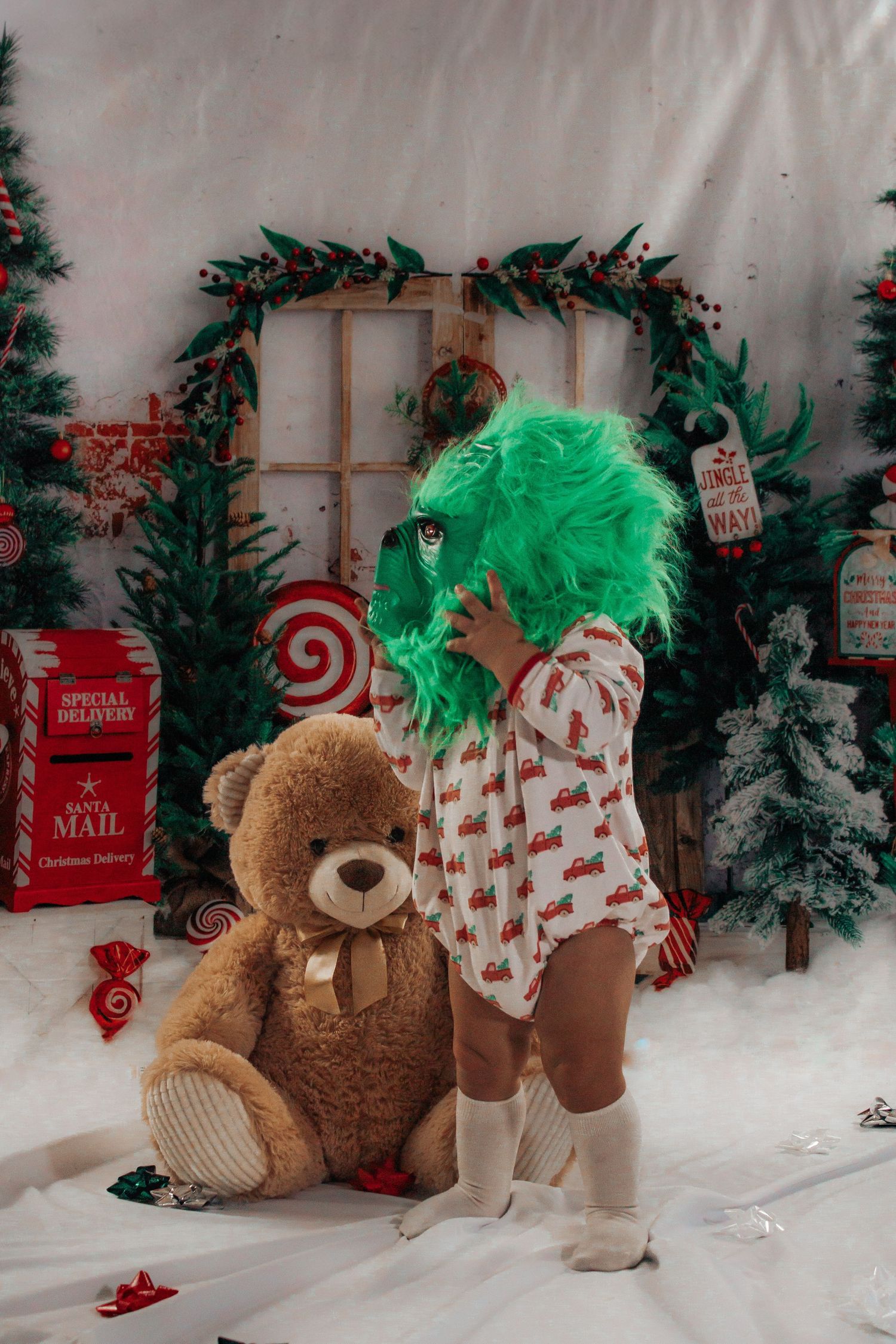 Child wearing Grinch mask hugs teddy bear in front of Christmas holiday backdrop.