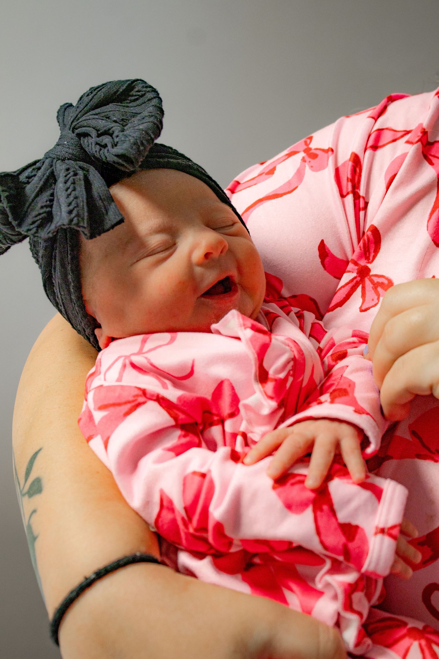 Newborn baby girl in pink outfit and bow headband yawning in mothers arms.