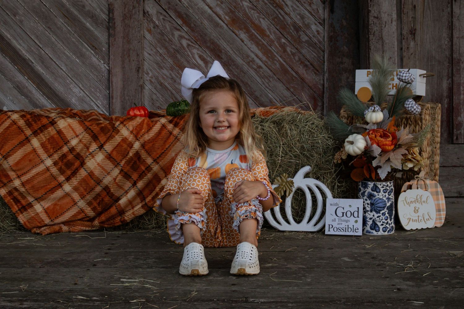 Smiling young girl in autumn outfit sitting on porch with fall harvest decorations and hay bales.