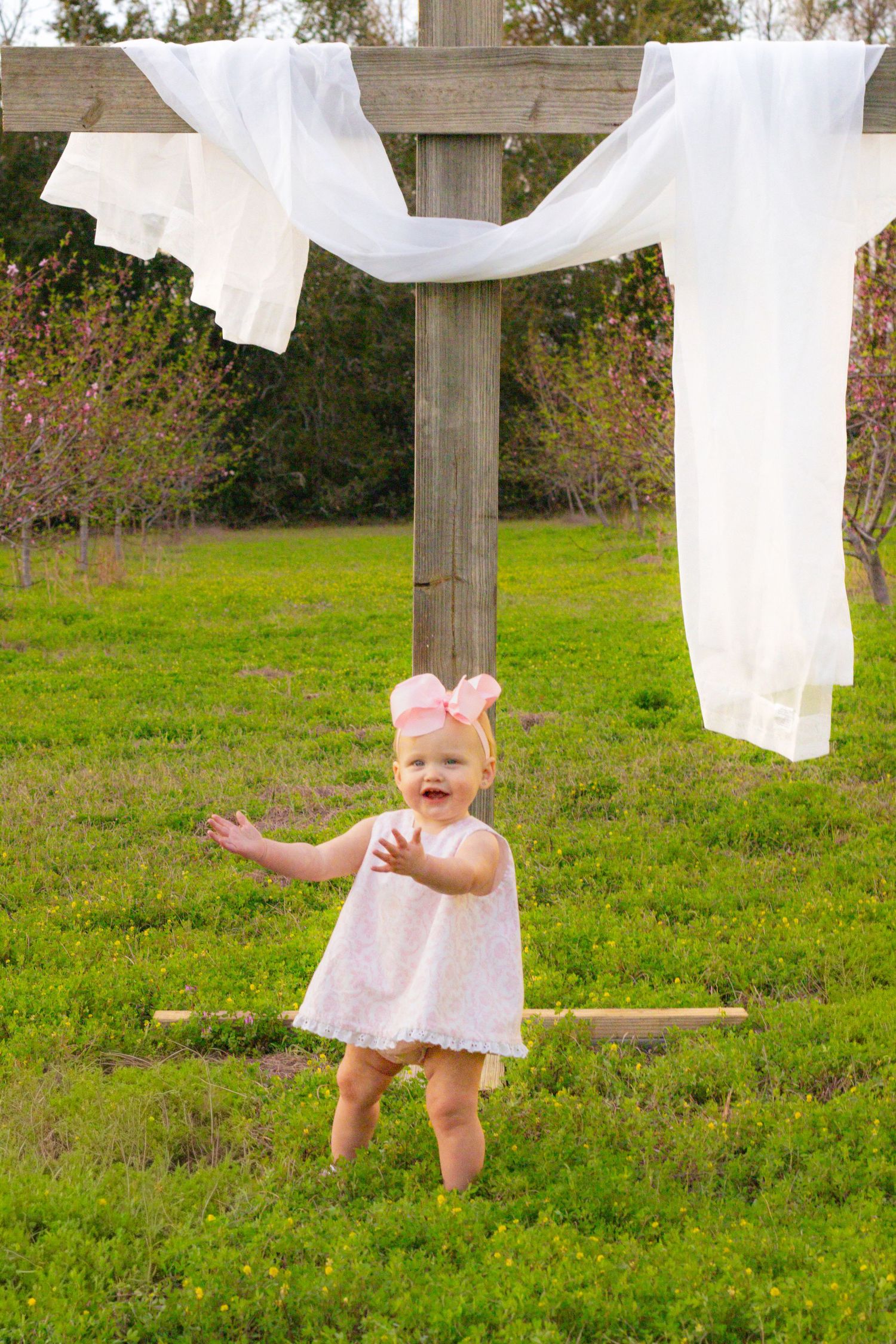 Joyful baby girl in white dress with pink bow reaches out while standing near a wooden cross outdoors.