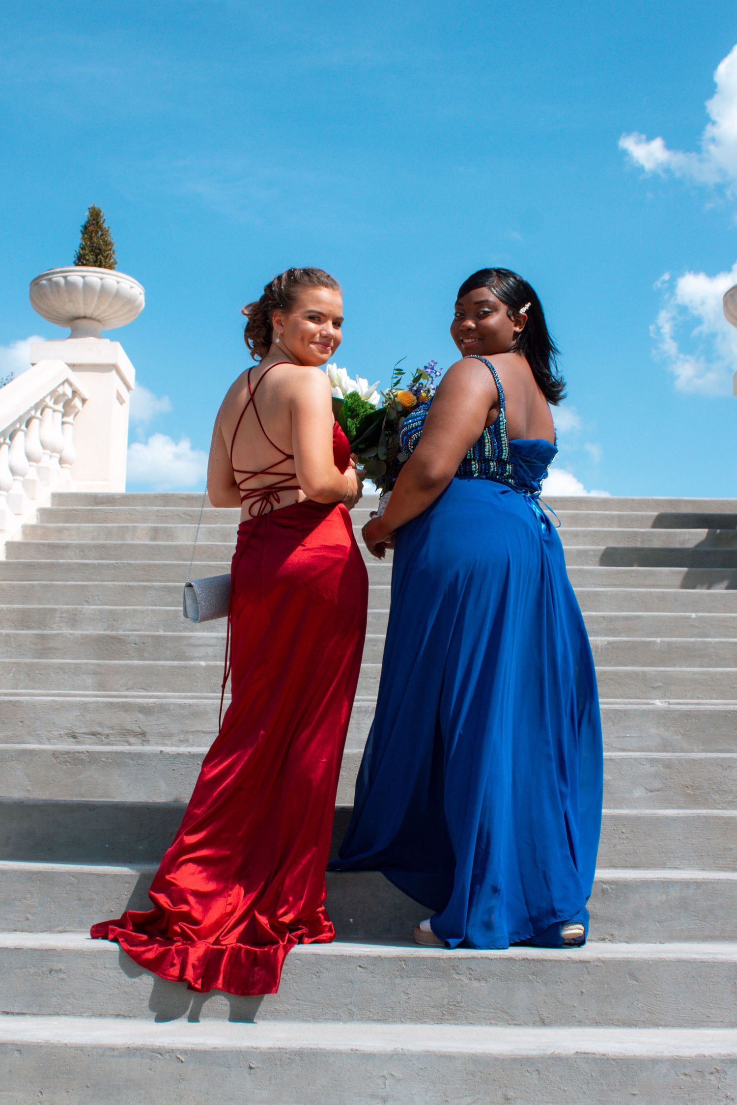 Two young women in red and blue formal gowns posing back-to-back on white marble stairs.