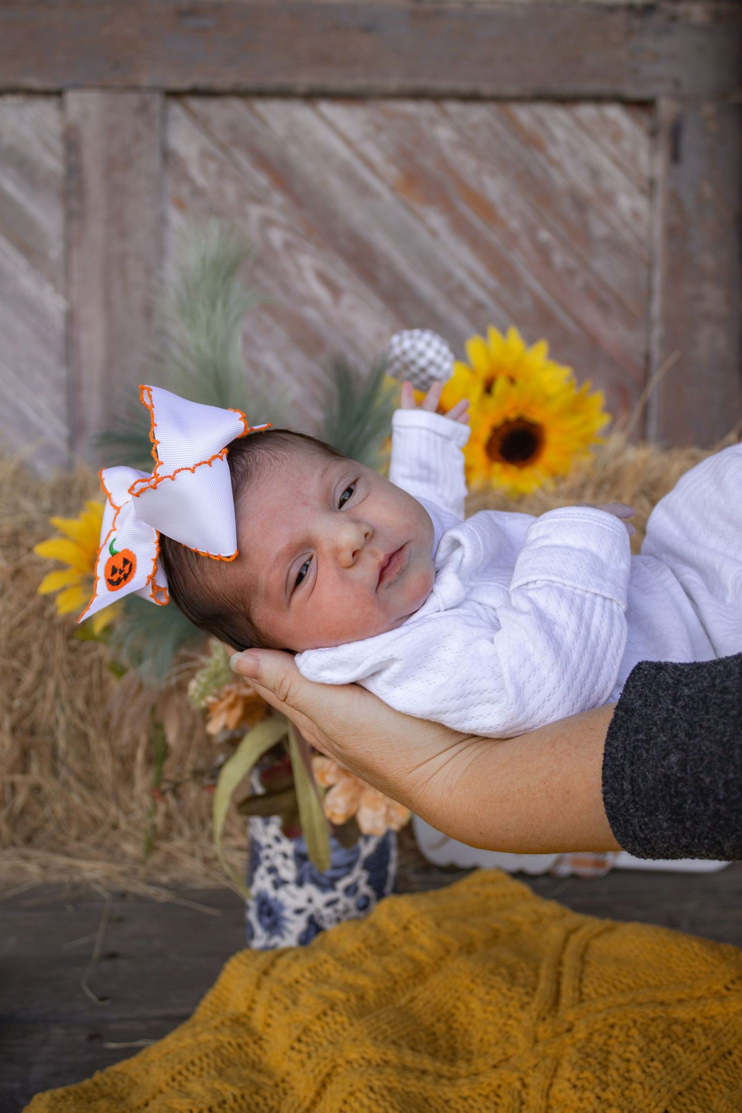 Newborn baby wearing a white bow headband lying on hay with sunflowers in a rustic fall-themed photo session.