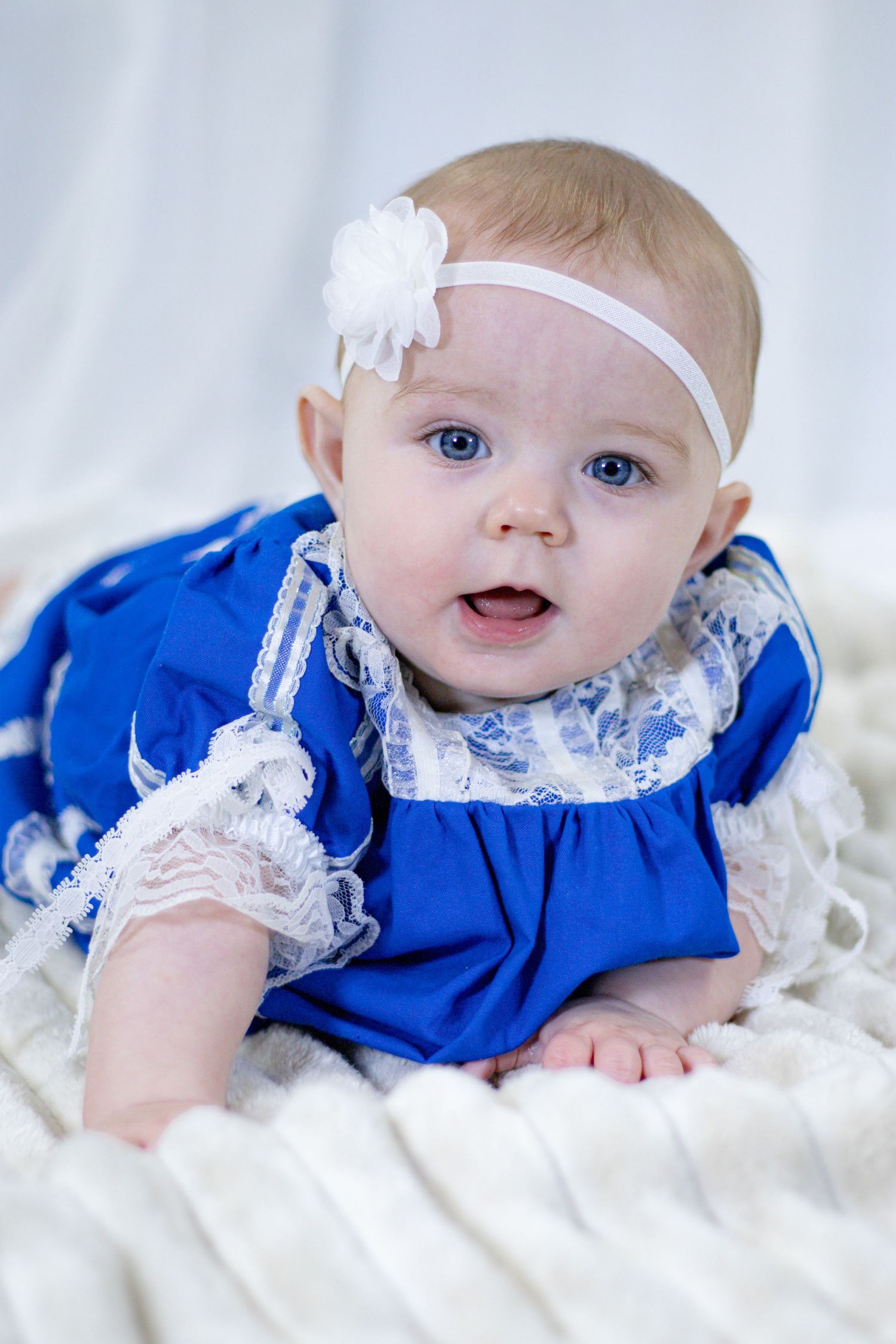 Blue-eyed baby girl in royal blue lace dress with white flower headband during tummy time portrait.