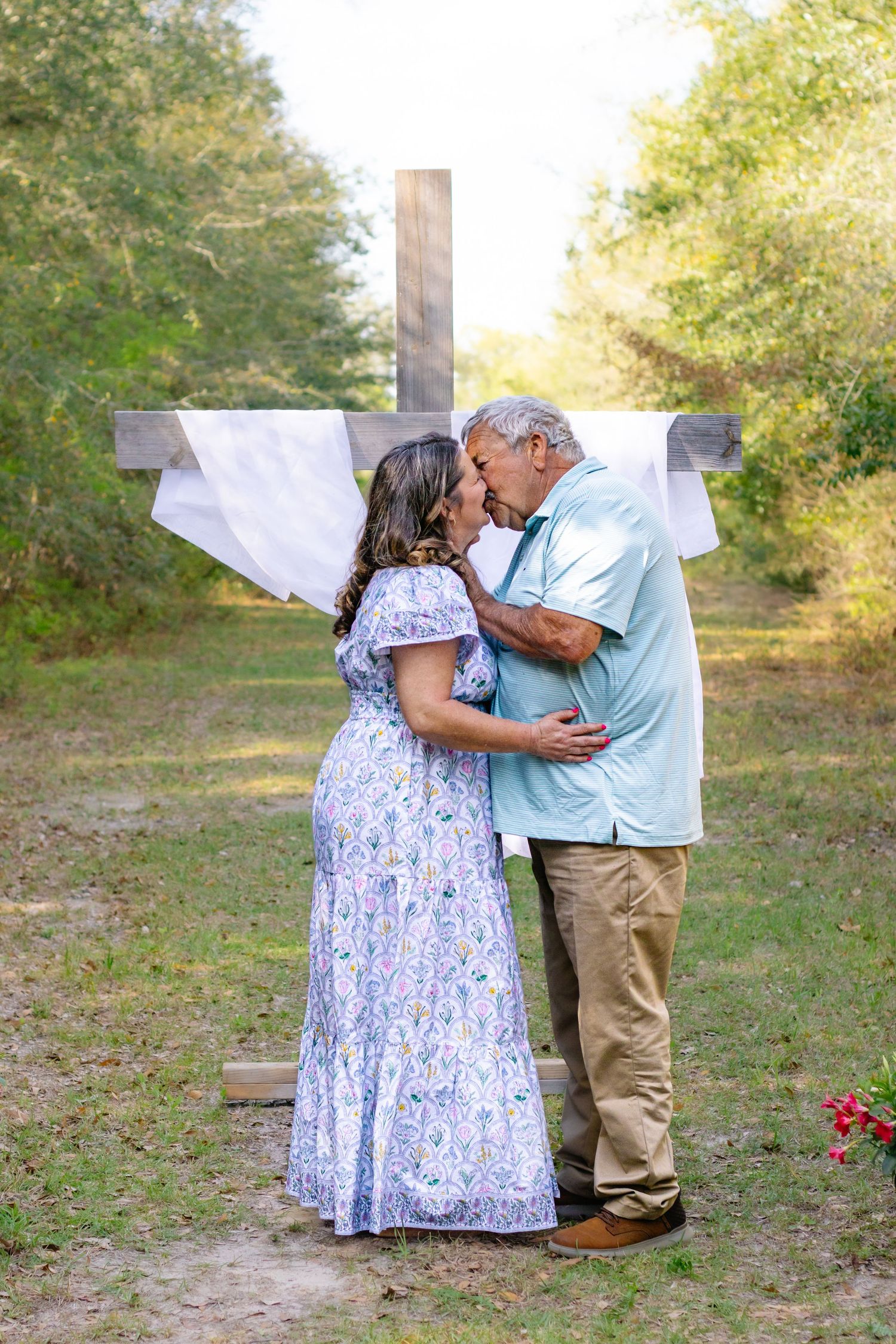 Couple sharing a romantic kiss in front of a rustic wooden cross in a natural outdoor setting.
