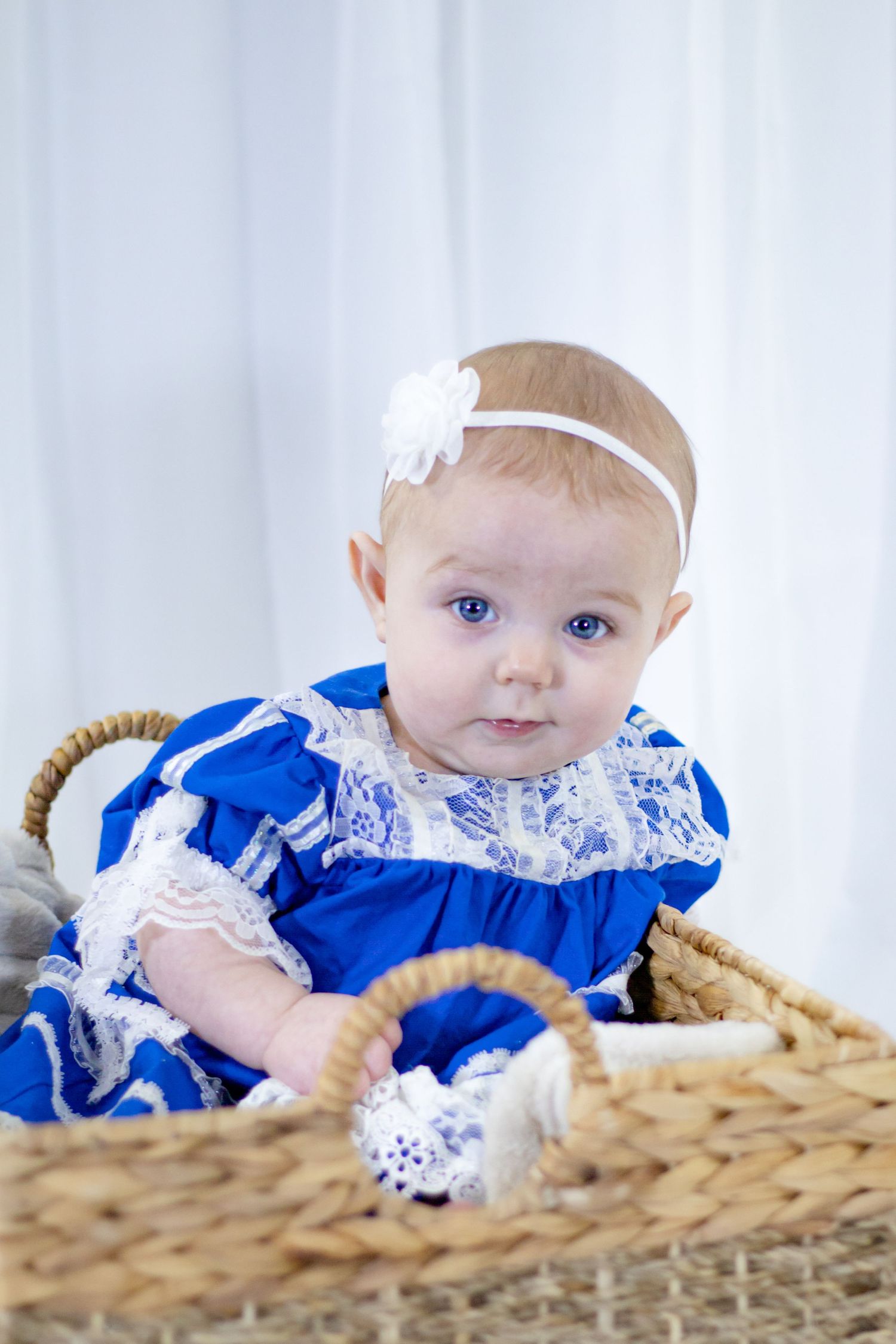 A baby girl in a blue lace dress and white floral headband sits in a wicker basket.