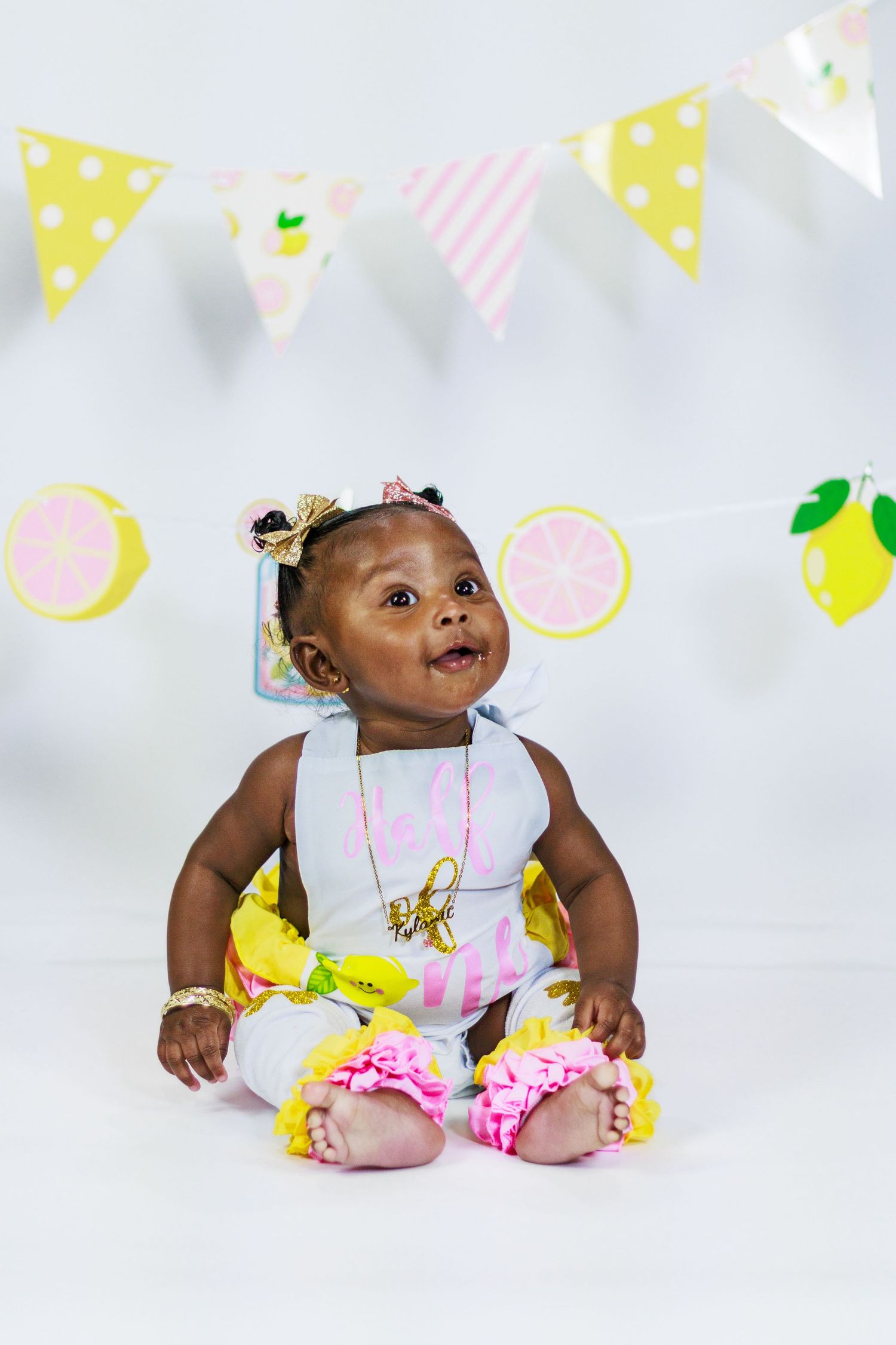 Baby girl in lemon themed outfit sitting near colorful citrus party banner decorations.