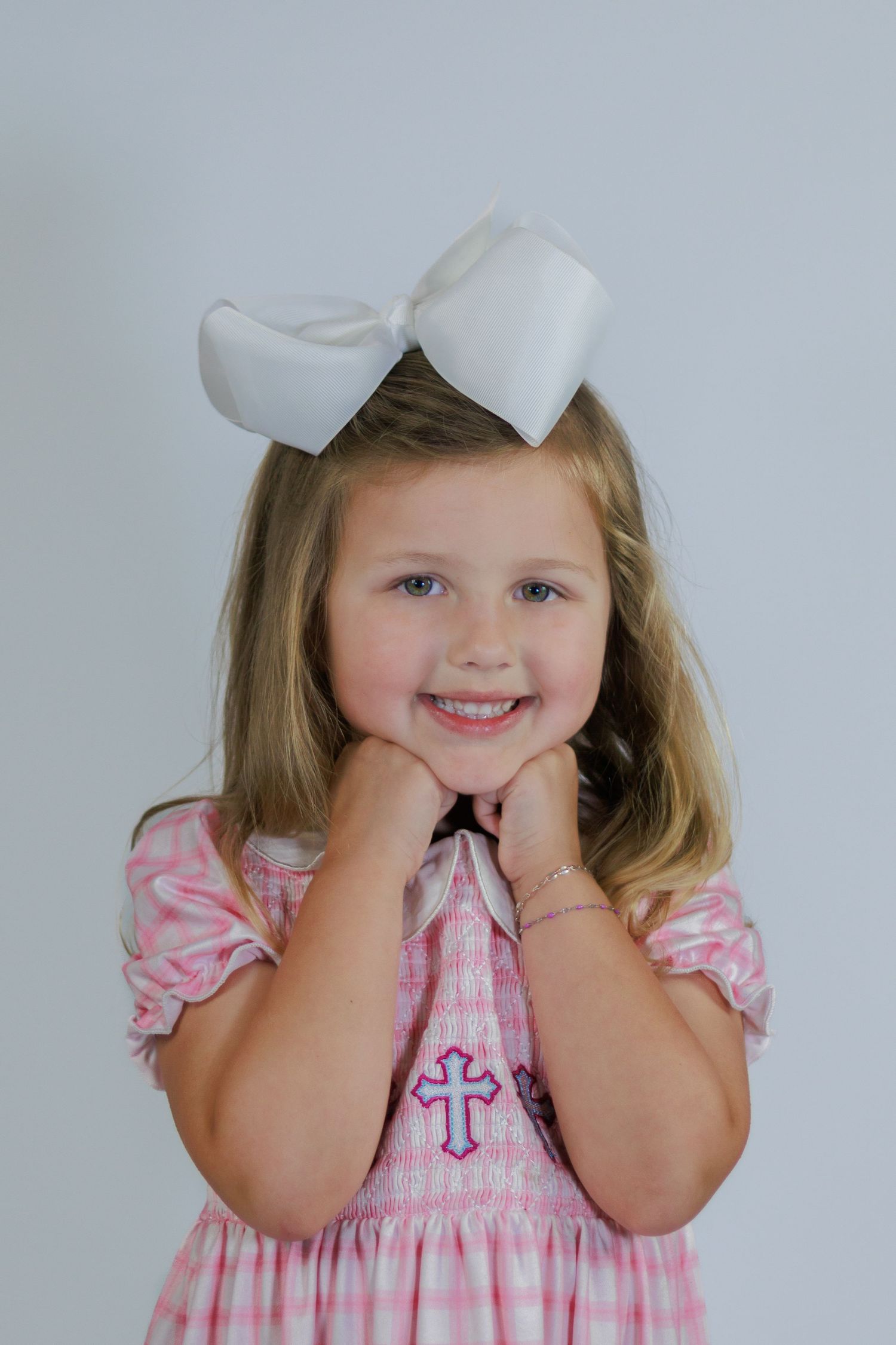 Smiling girl in pink smocked dress with cross embroidery and white hair bow poses cheerfully.