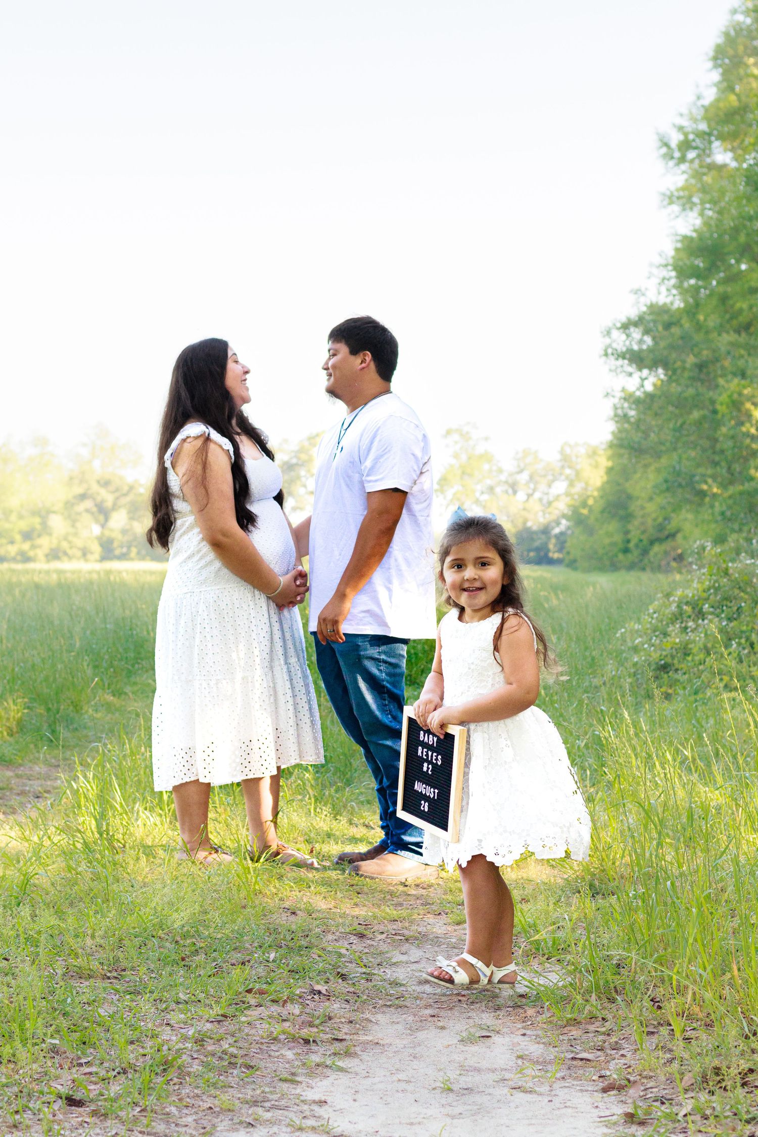 Pregnant woman in white dress holds hands with partner while daughter holds pregnancy announcement sign outdoors.