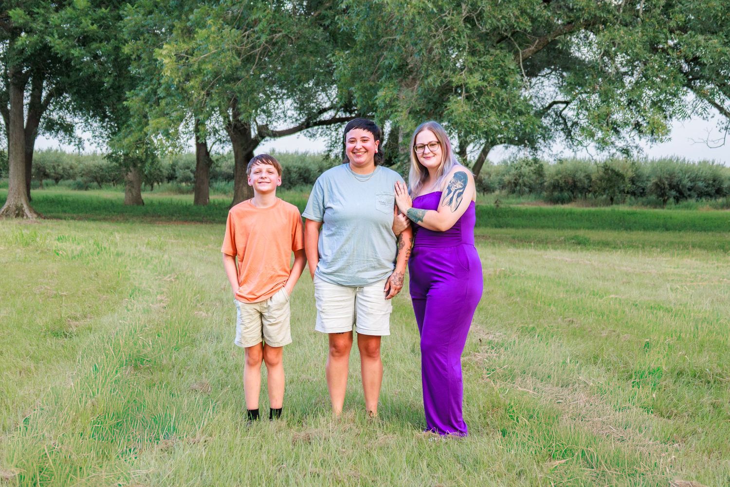 Three people smiling outdoors on a green grass path surrounded by trees on a sunny day.
