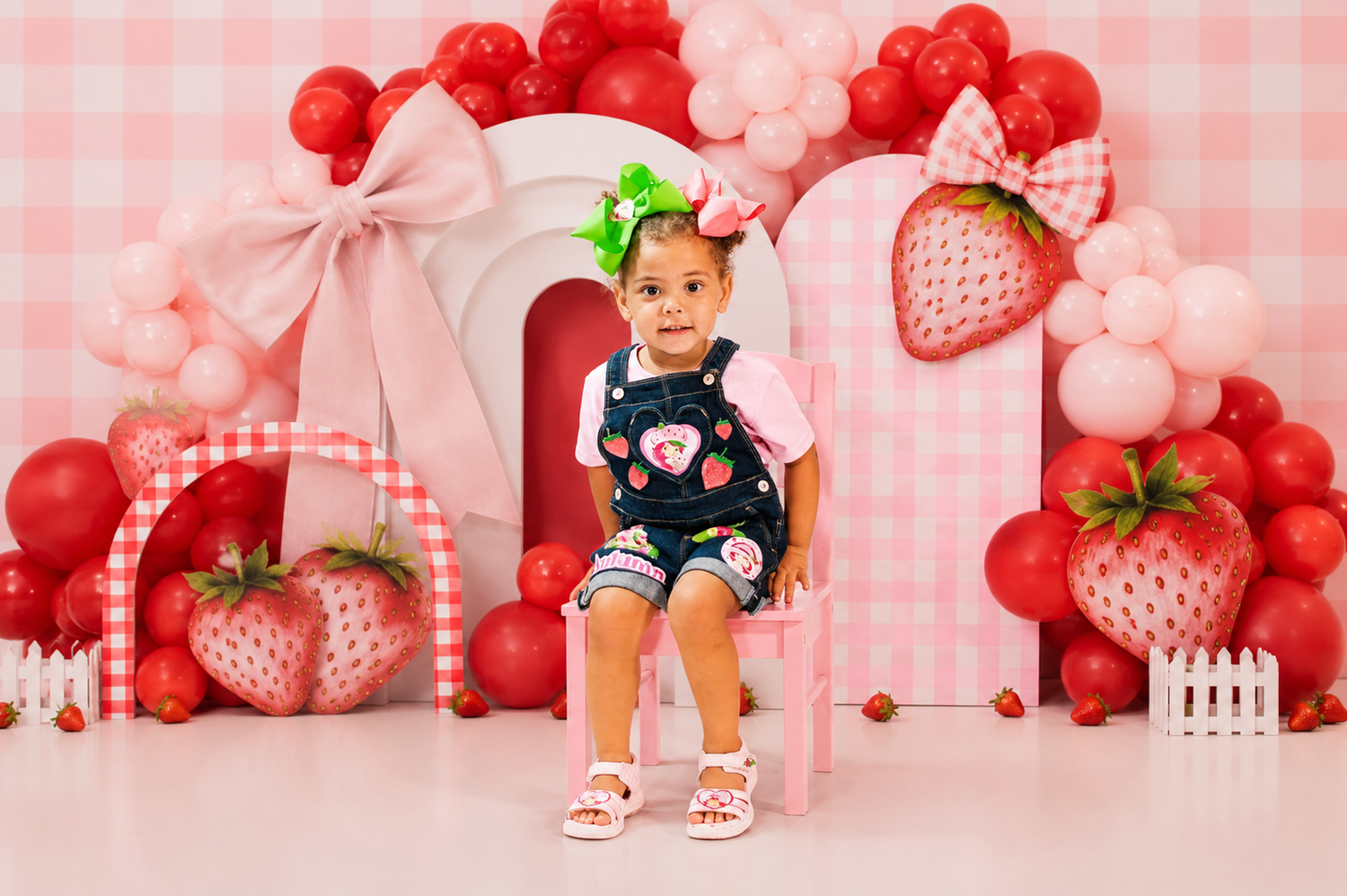 Toddler girl in denim overalls sits in strawberry-themed photo backdrop with balloons and decorations.