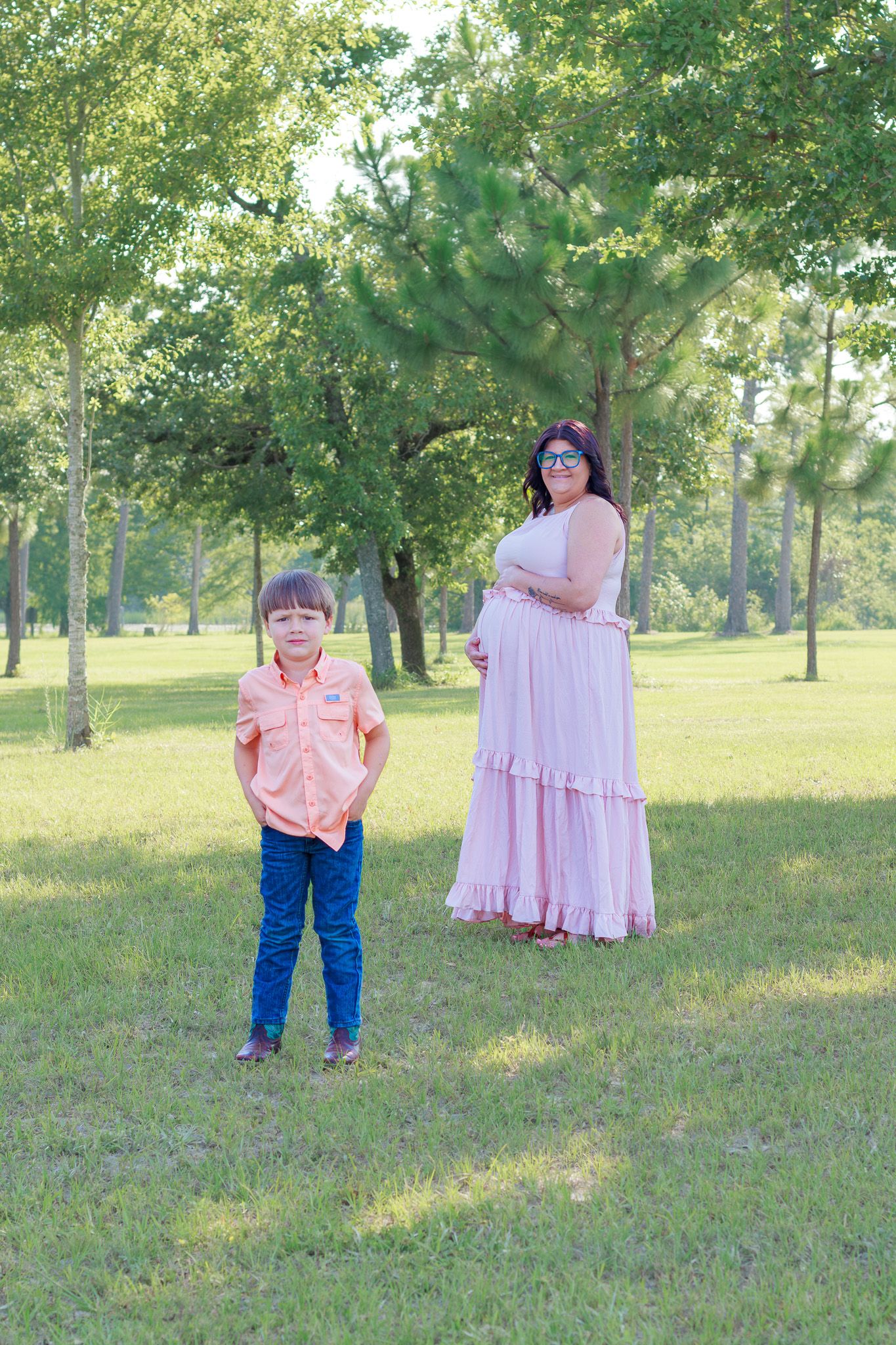 A pregnant woman in a pink dress stands with a young boy in an orange shirt in a sunny green park.