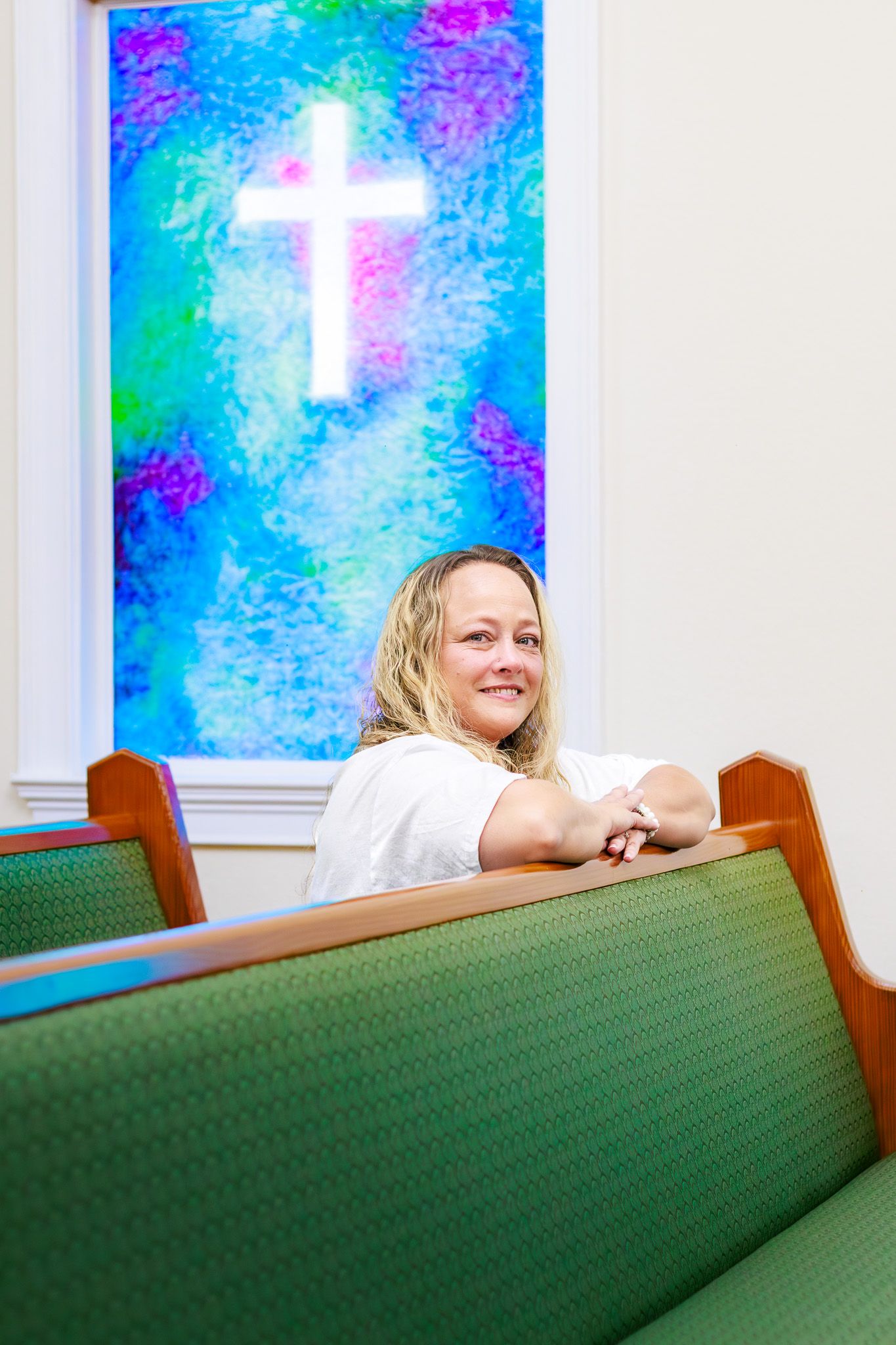 Woman smiling in church pew with colorful stained glass window and glowing white cross behind her.