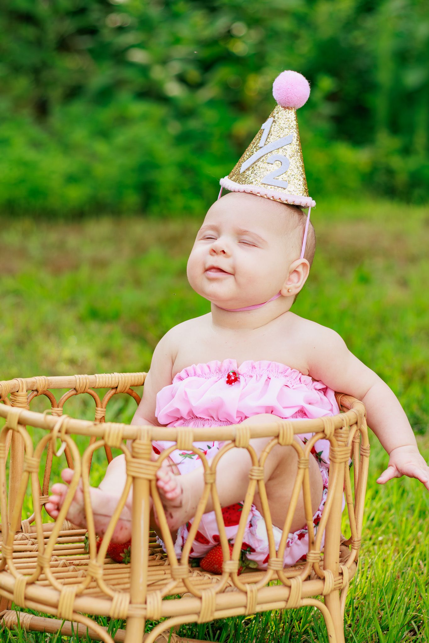 Cute baby in pink ruffles and gold birthday hat sits smiling in wicker basket on green grass.