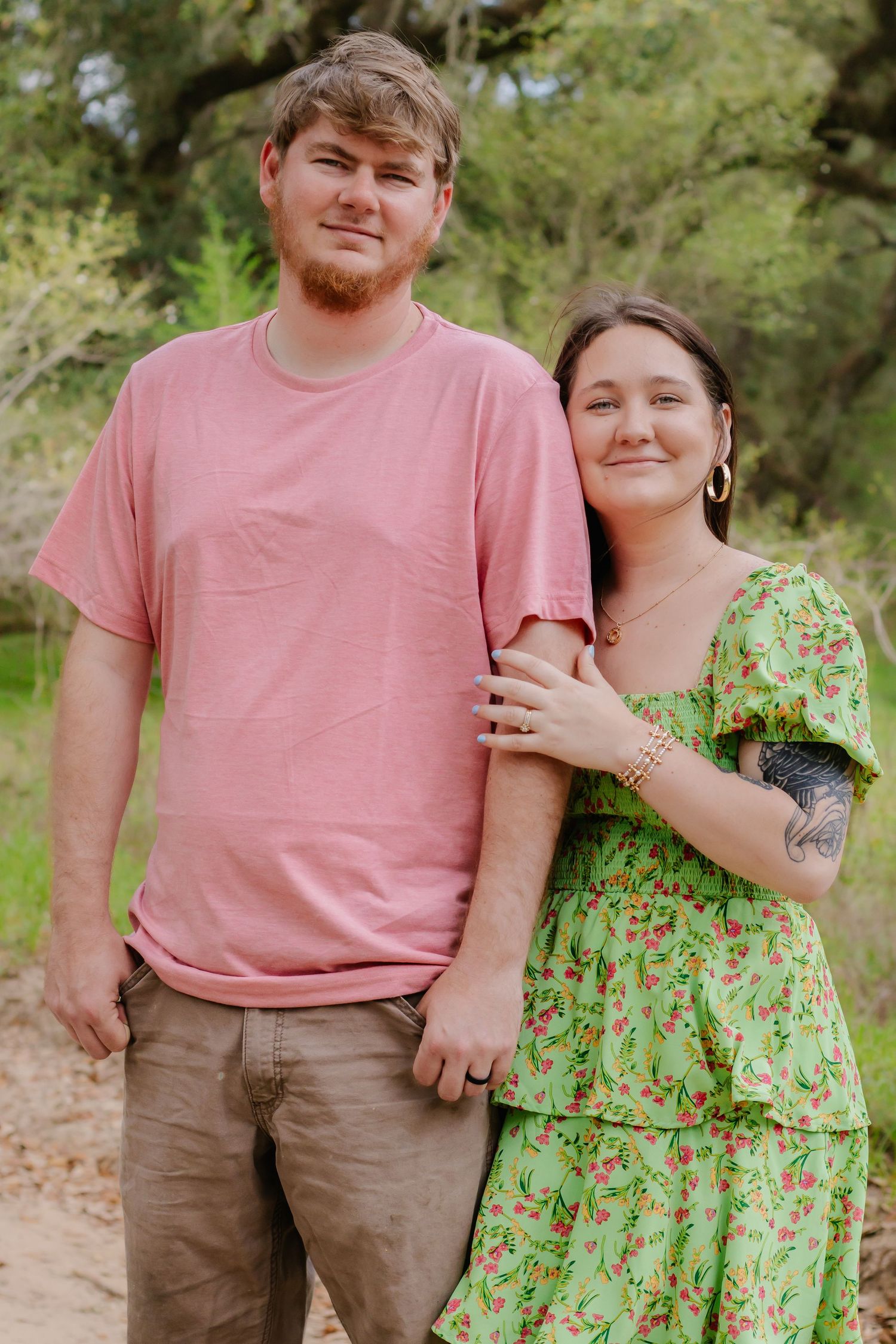 Young couple posing outdoors, man in pink shirt and woman in green floral dress.