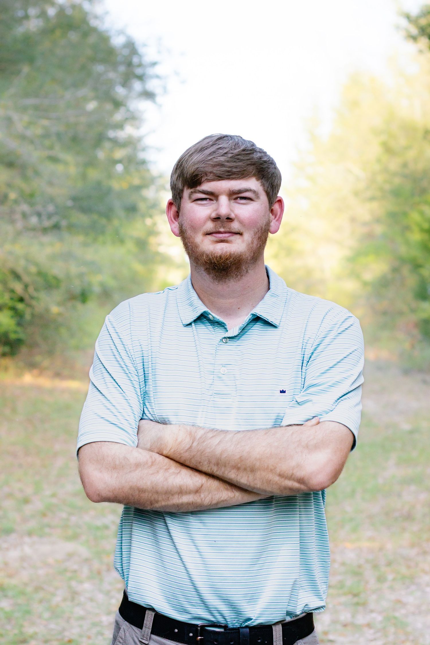 Young bearded man in a light blue polo shirt standing outdoors with arms crossed.