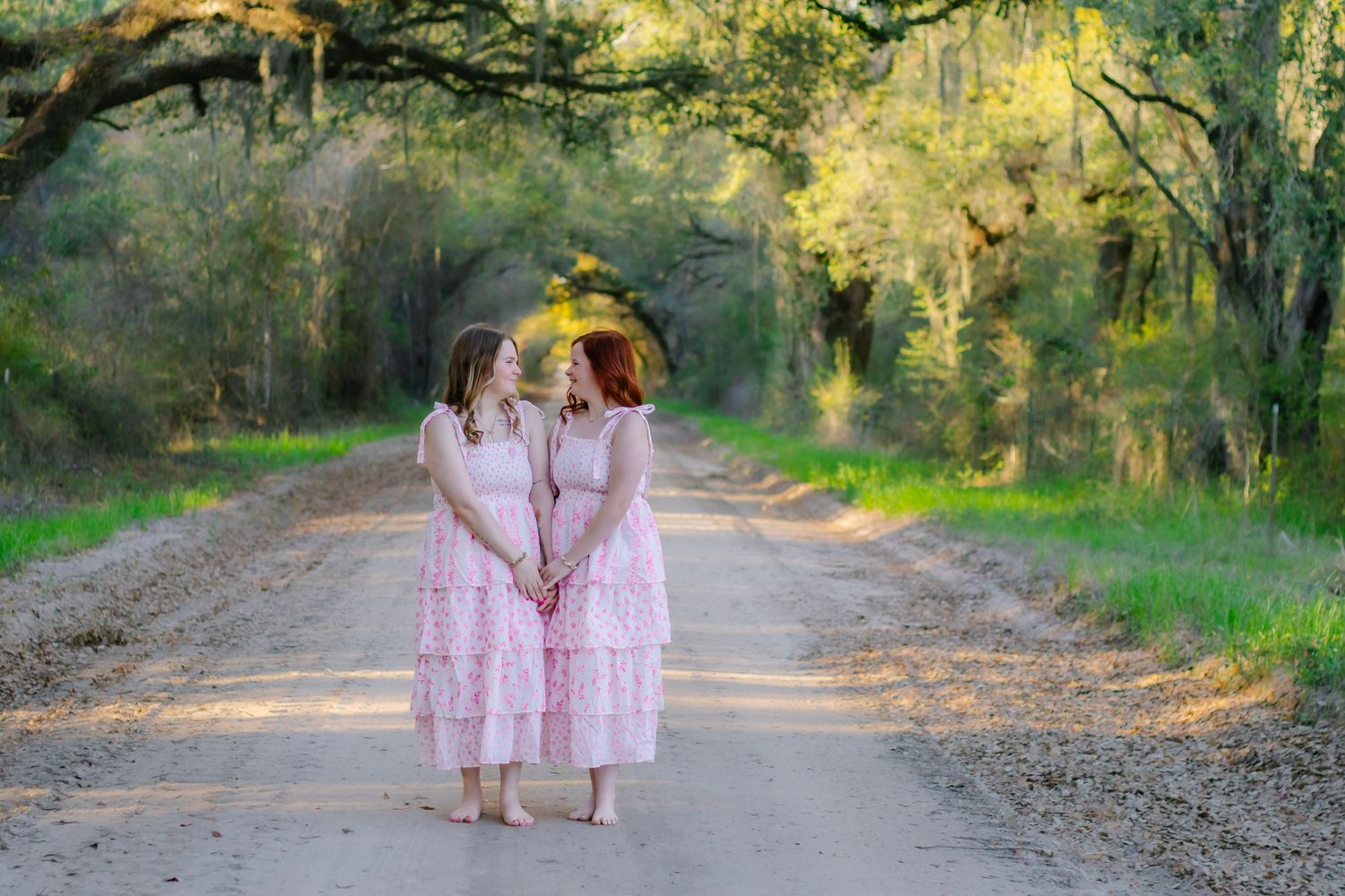 Two girls in matching pink floral dresses hold hands on a scenic tree-lined dirt road.
