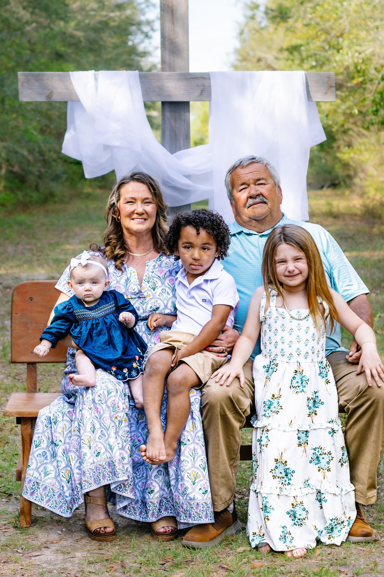 Grandparents seated on bench with three grandchildren before a white-draped wooden cross in outdoor setting.